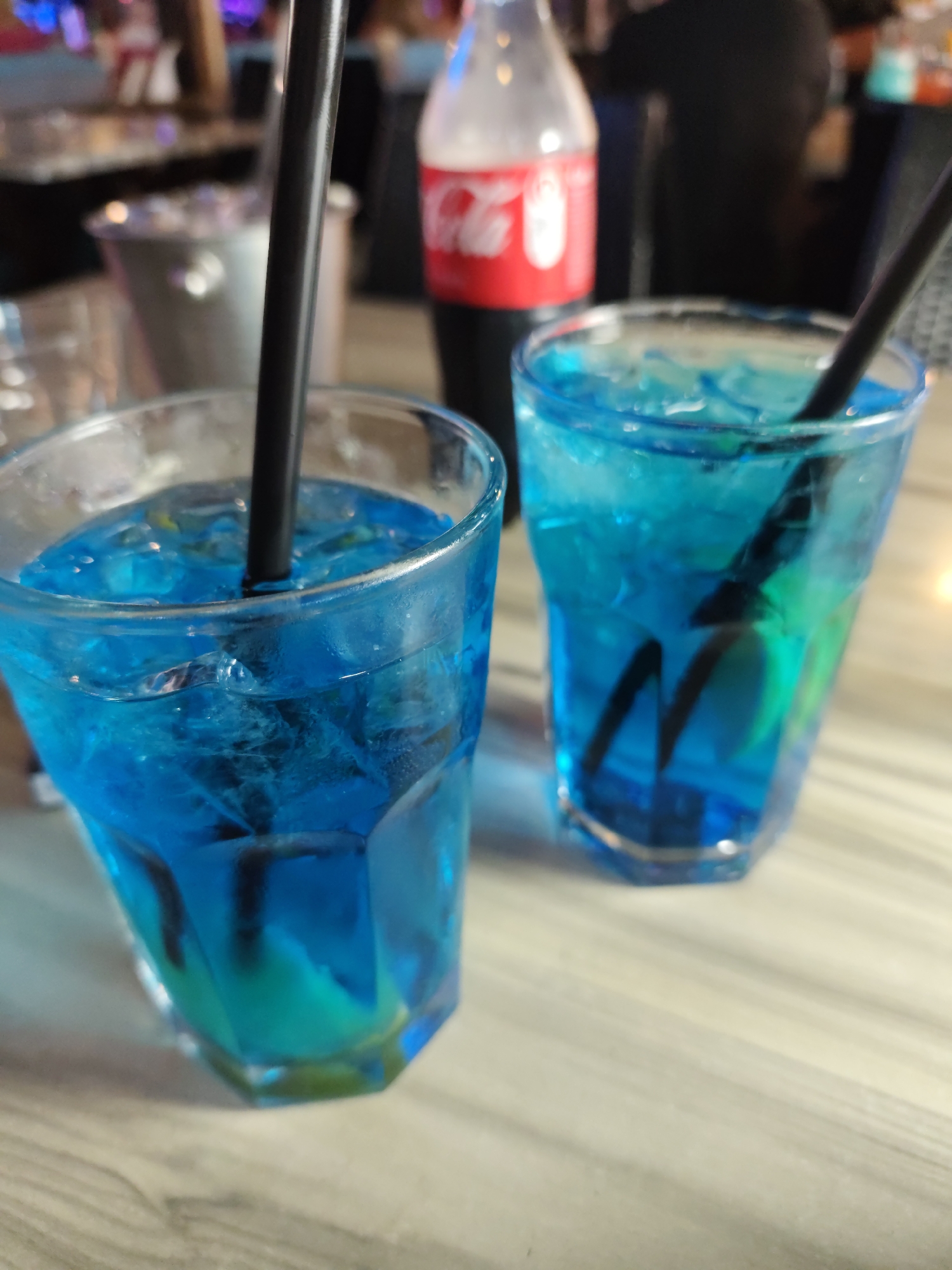Two glasses of blue liquid with ice and black straws are on a wooden table. A Coca-Cola bottle is in the background. The scene appears to be in Nesebar, Bulgaria, likely a bar or restaurant.