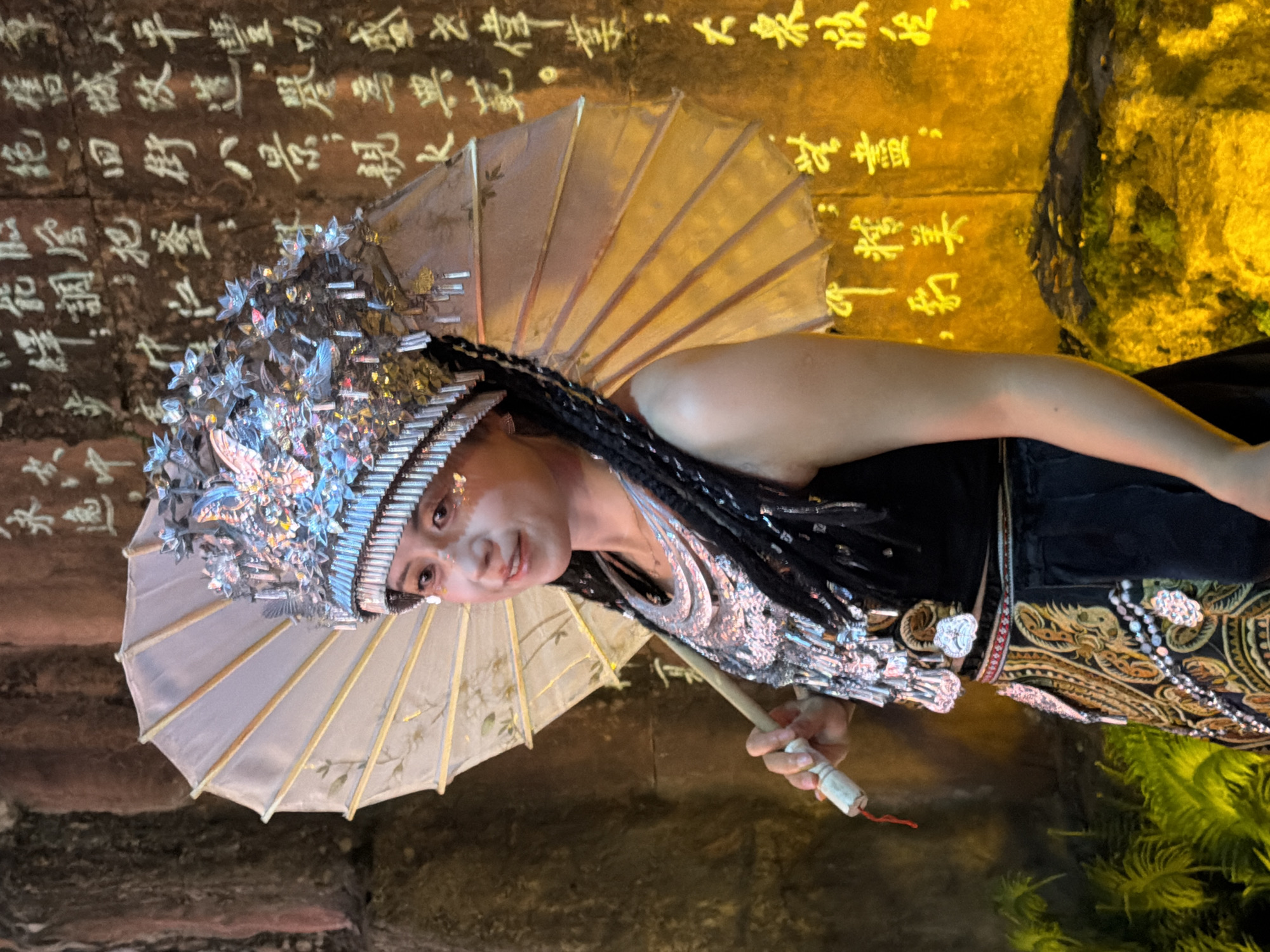 A person dressed in a traditional Chinese costume is performing in what appears to be a historical or cultural setting. The individual wears an elaborate headdress adorned with silver ornaments and what looks like flowers, and holds a small, light-colored parasol. Their outfit is detailed, featuring a black top with silver embellishments and a richly patterned skirt or sash with gold and black designs. The performer has dark hair styled in braids.
The background consists of a stone wall with inscriptions in Chinese characters. The lighting suggests it might be evening or indoors, with spotlights highlighting the wall and the performer. To the right, there's a rocky outcrop with some greenery. The overall atmosphere seems to be one of cultural performance or a historical reenactment, possibly during a festival or special event. The Chinese text on the wall likely adds to the historical or cultural context of the performance.