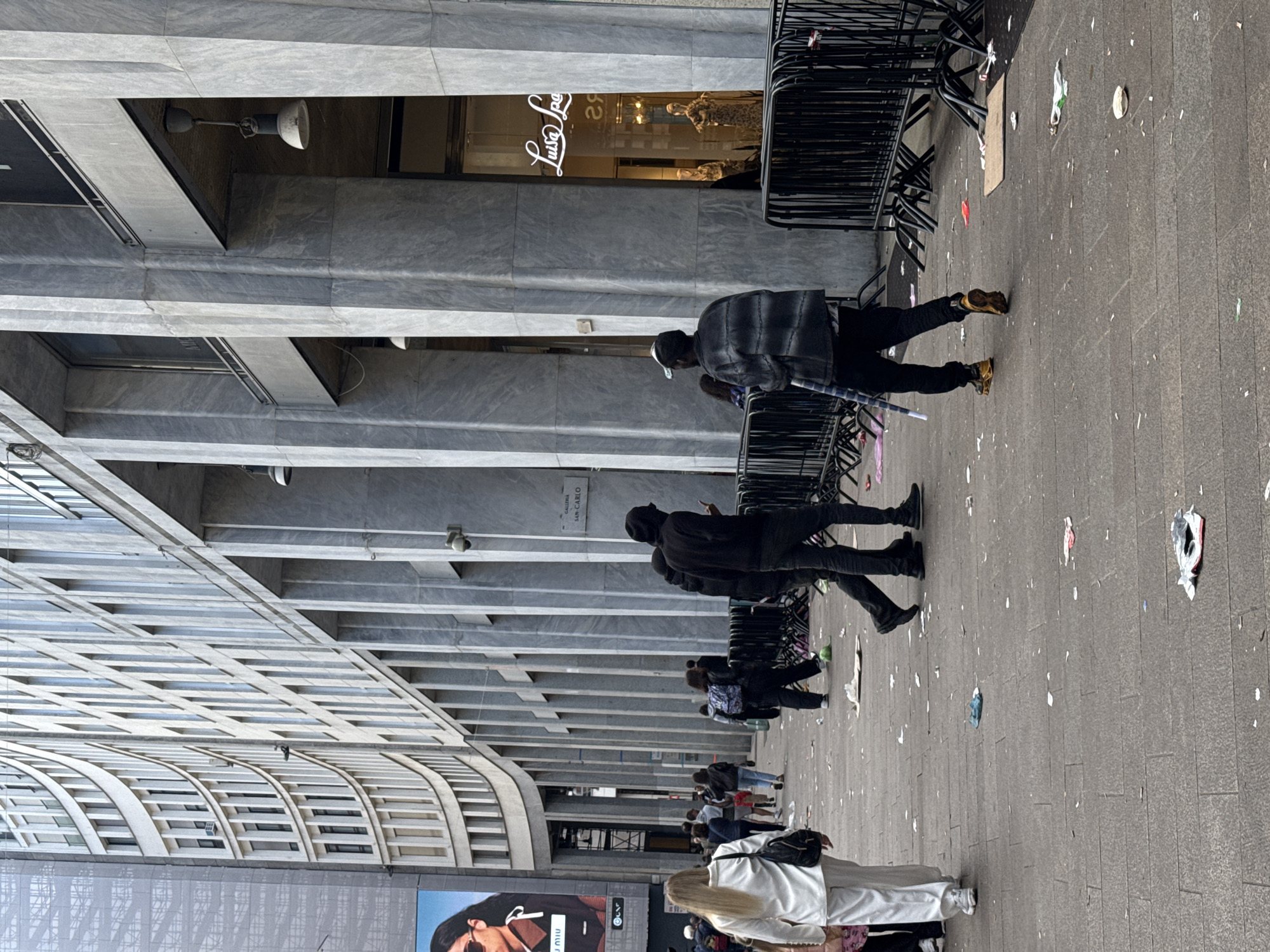 The image depicts a street scene in Milan, Italy. A group of people is walking along a paved sidewalk next to a building with a marble facade. The sidewalk is littered with debris. Metal crowd control barriers are present. A store with a "Luisa Spagnoli" sign is visible in the background. A billboard featuring a "Miu Miu" advertisement is also visible. The architecture indicates a central, possibly upscale area of the city.