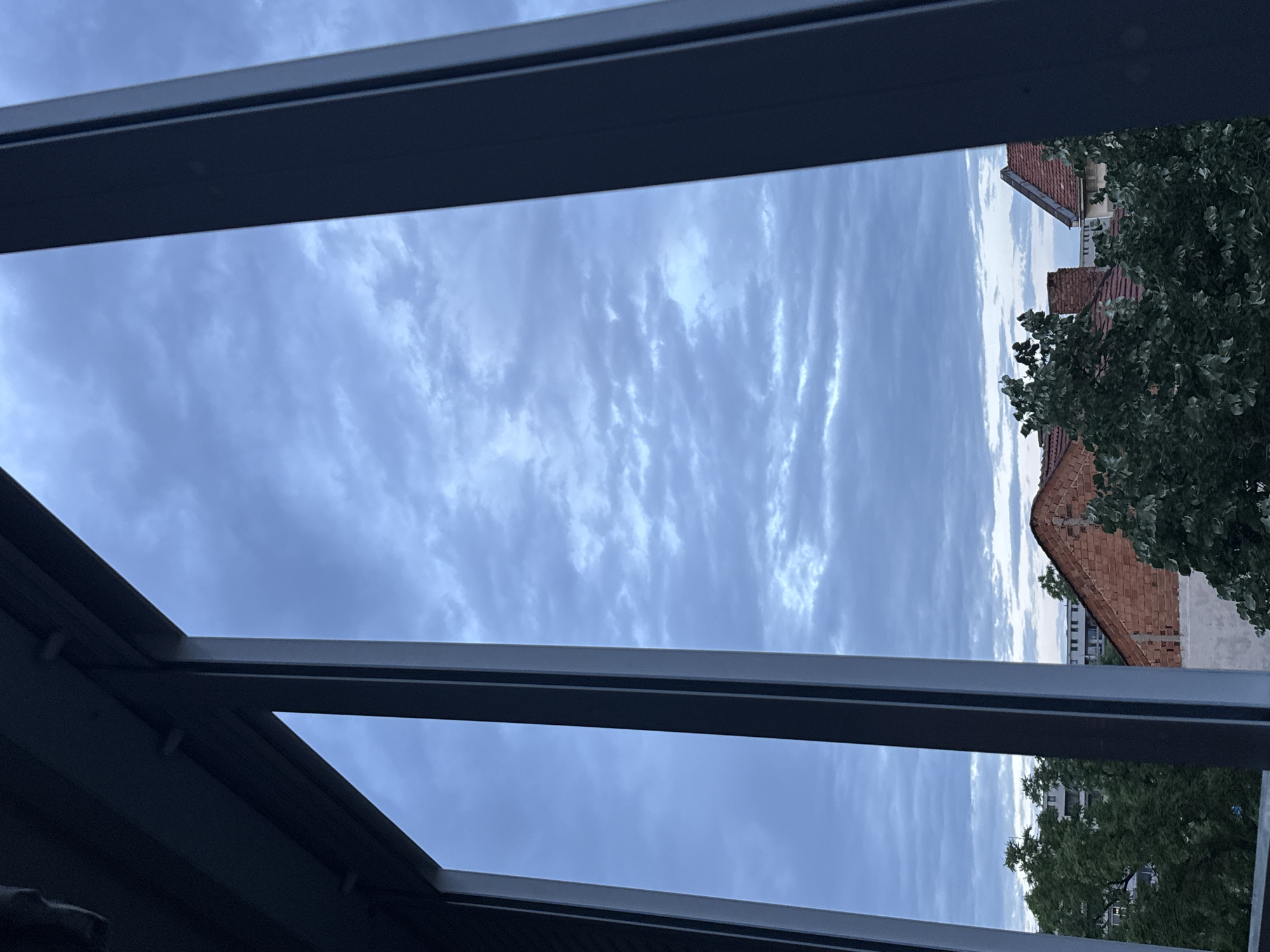 This image presents an outdoor scene viewed through a dark, possibly metallic, architectural frame, suggesting a window, skylight, or modern canopy. The dominant subject is the sky, filled with an extensive array of clouds. These clouds form distinct, elongated patterns and ripples across the blue-gray expanse, characteristic of altocumulus or cirrocumulus formations, creating a textured and dynamic overhead view.
In the right portion of the frame, beneath the cloudy sky, an urban or suburban landscape is visible. Several buildings with traditional red-tiled roofs and brick facades are present, along with a prominent brick chimney. Mature green trees with dense foliage partially obscure some of these structures. The architectural style, particularly the red-tiled roofs, is consistent with buildings found in European cities, aligning with the provided context of Plovdiv, Bulgaria.
The time of day appears to be daytime, but the absence of direct sunlight and the pervasive cloud cover indicate an overcast day, or possibly early morning or late afternoon with diffused light. No people or specific events are taking place; the image captures a static, contemplative view of the sky and rooftops. The weather is cloudy but not stormy, with the clouds forming an artistic backdrop. There is no visible text in the image.