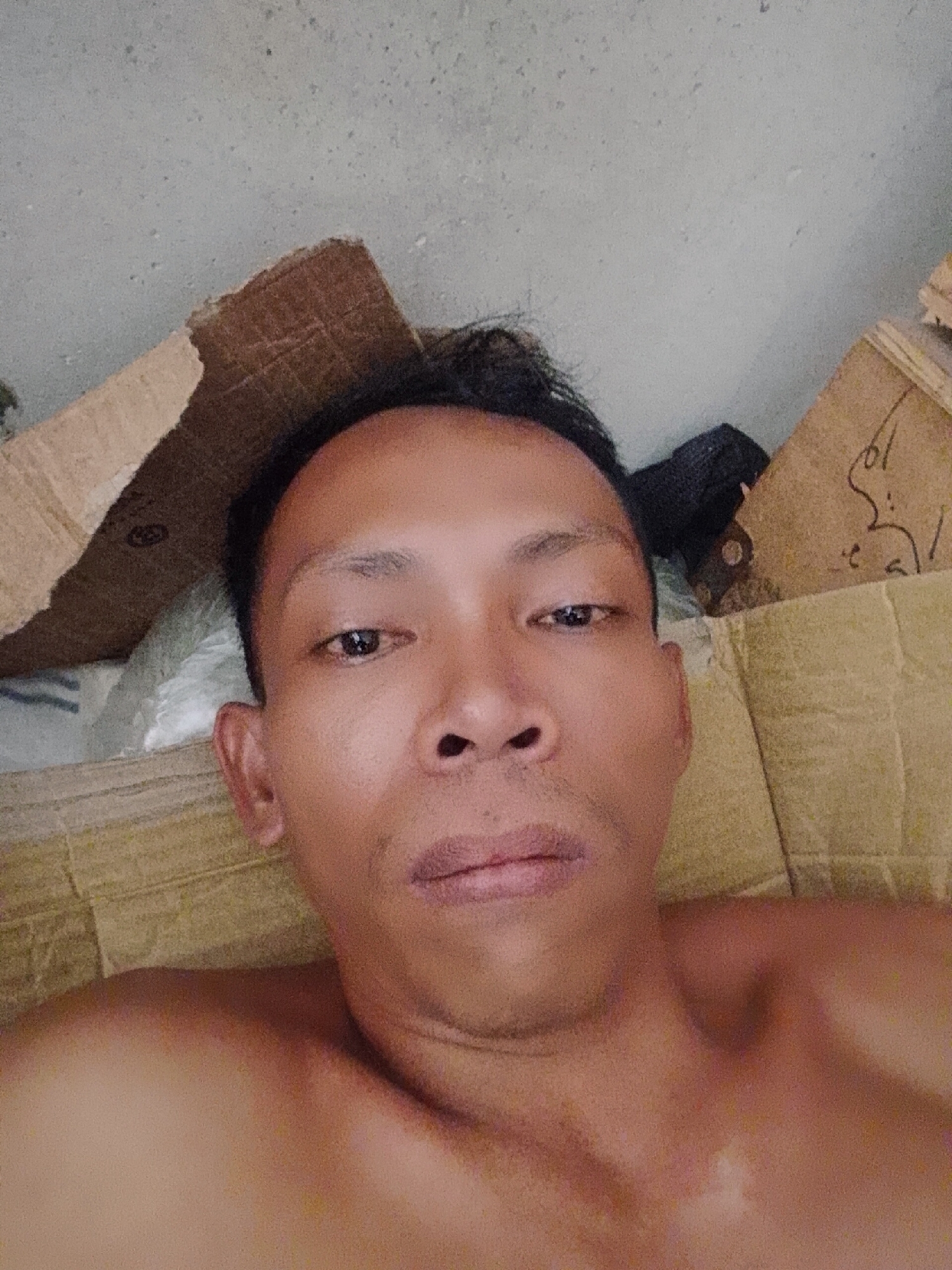 The image shows a medium close-up, selfie-style shot of a bare-chested man with short black hair. He has brown eyes and appears to be lying down on a surface with a textured, yellow or beige material. In the background, there are pieces of brown cardboard and what seems to be a dark-colored object. There is no discernible information about a location or setting within Bukit Ketok, Indonesia. The provided information does not permit the determination of the location, actions, or interactions.