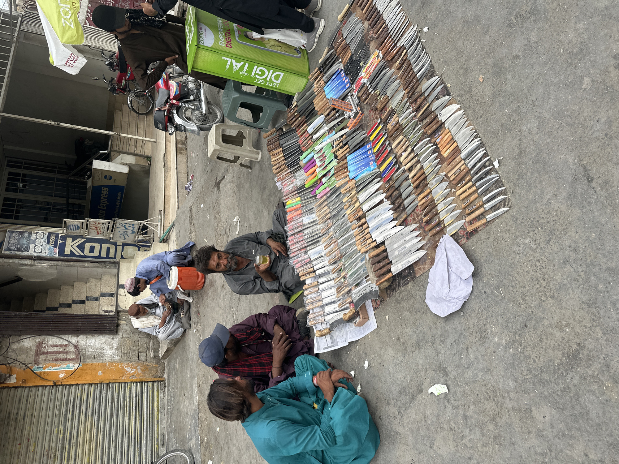 The image shows a street scene in Quetta City Tehsil, Pakistan. Three men are sitting on the ground next to a large display of knives laid out on the pavement. The knives vary in size and style, many still in packaging. One of the men is holding a glass. Beside the display is a green plastic crate with the words "DIGITAL" on it. Behind the men and the knife display is a building with a partially closed metal shutter and shop signs in Urdu and English, including one that reads "Photo Express". A couple of other men are standing near the Photo Express store. One of them is carrying what appears to be an orange cooler or container. Beyond the first building is a street with parked motorcycles. The sky is overcast.