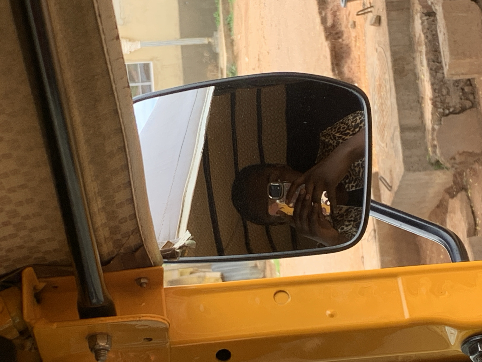 The image displays a reflection in a black-framed side-view mirror of a bright yellow commercial tricycle, originating from New Stadium, Nigeria. Inside the mirror's reflection, an individual with dark skin and short dark hair is visible, holding a smartphone with both hands directly towards themselves, indicative of taking a selfie. The smartphone is encased in a light-colored, patterned case. The individual is wearing a garment with a distinct animal print, possibly leopard. The reflected interior of the tricycle shows a brown, textured ceiling panel and several dark horizontal structural elements.
The external environment visible around and through the mirror reveals an unpaved, reddish-brown dirt ground. In the background, a light-yellow building with a white-framed window and a white vertical column or pipe is partially visible. Concrete structures, resembling steps or a raised platform, are present on the right side of the foreground, accompanied by scattered rocks and debris on the ground.