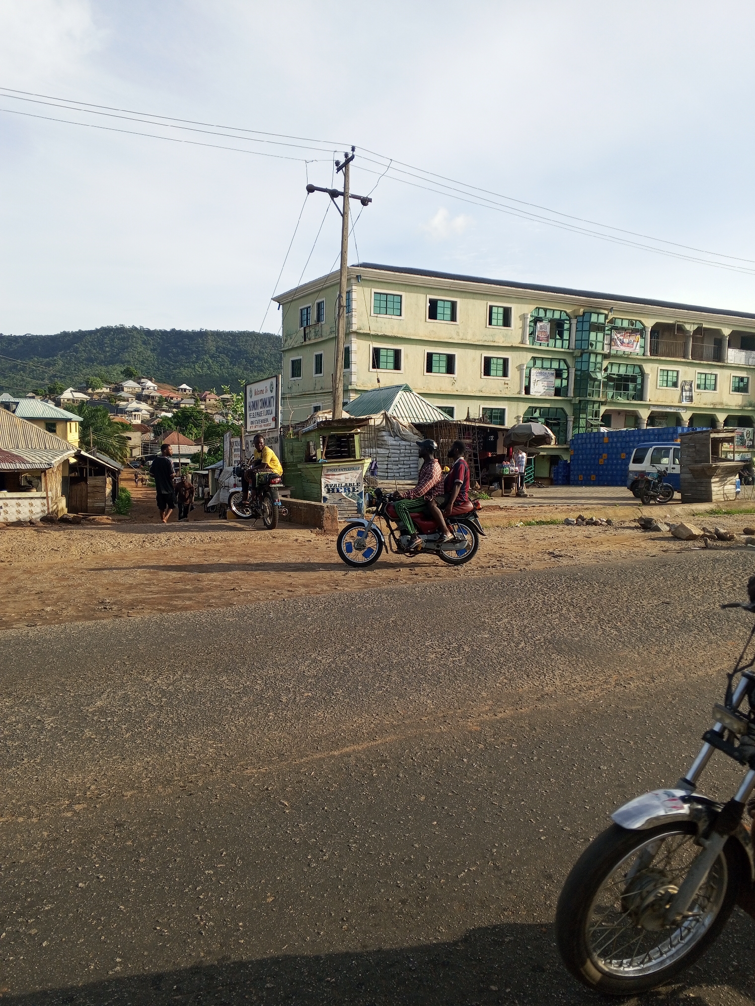 The image shows a street scene in Lokoja, Nigeria. A paved road occupies the foreground. Two motorcycles are prominent: one in motion with two riders and another partially visible on the right edge. In the background, there are several informal structures, including small shops and homes, typical of a roadside commercial area. A three-story building, possibly a hotel or commercial property, is the largest structure visible. Mountains rise in the distance. Overhead power lines are present. People are milling around in the midground, near the shops. A sign reading "ALPHOMA COMMUNITY" is visible.