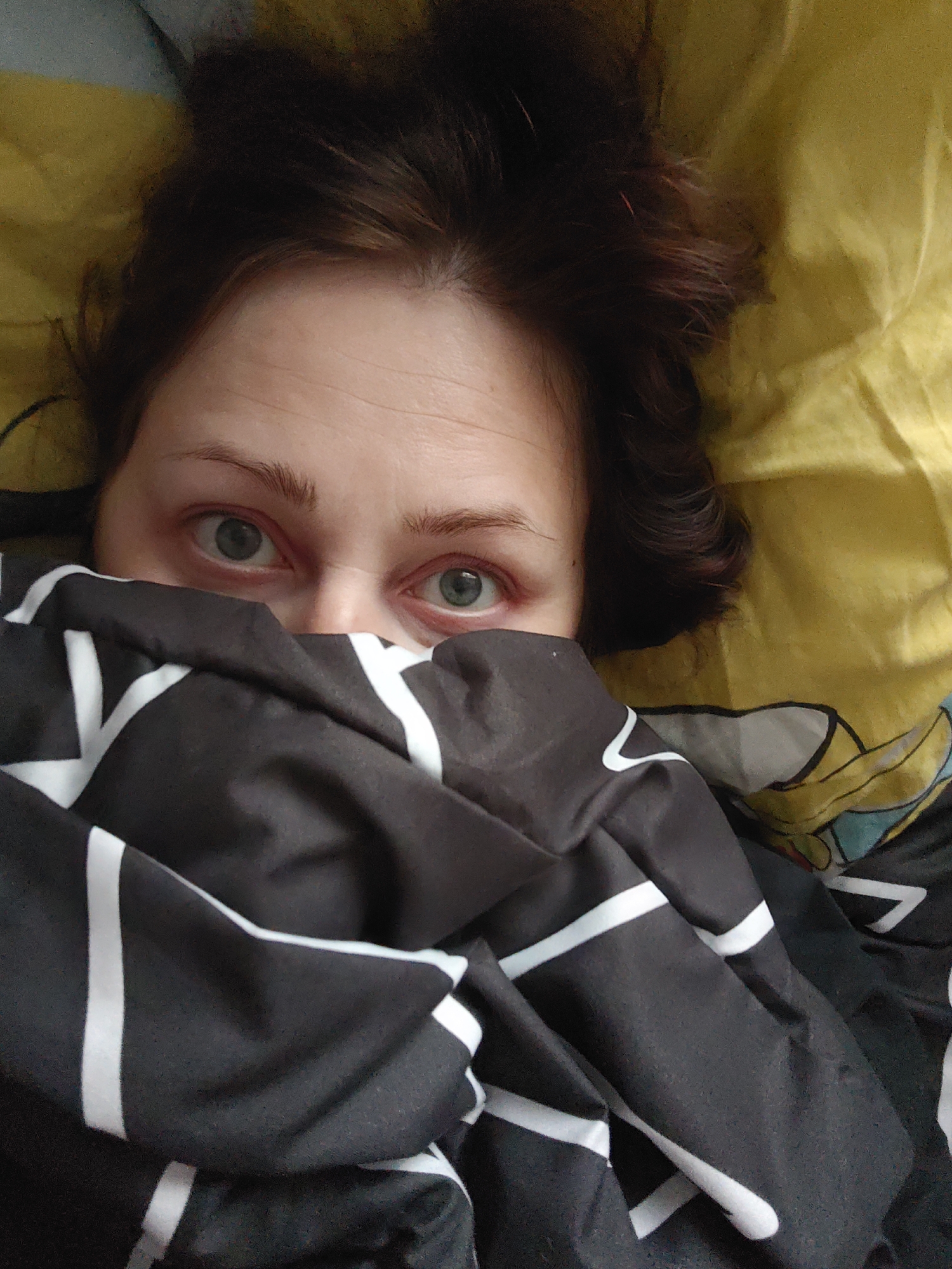 A selfie-style, close-up shot depicts a woman partially covered by a black and white geometric patterned blanket, lying on a yellow patterned bedspread. Only the upper portion of her face, from her eyebrows to the bridge of her nose, is visible. She has light skin, brown hair, and grey eyes. The background suggests an indoor environment, likely a bedroom. The location is specified as Марковский, Russia.