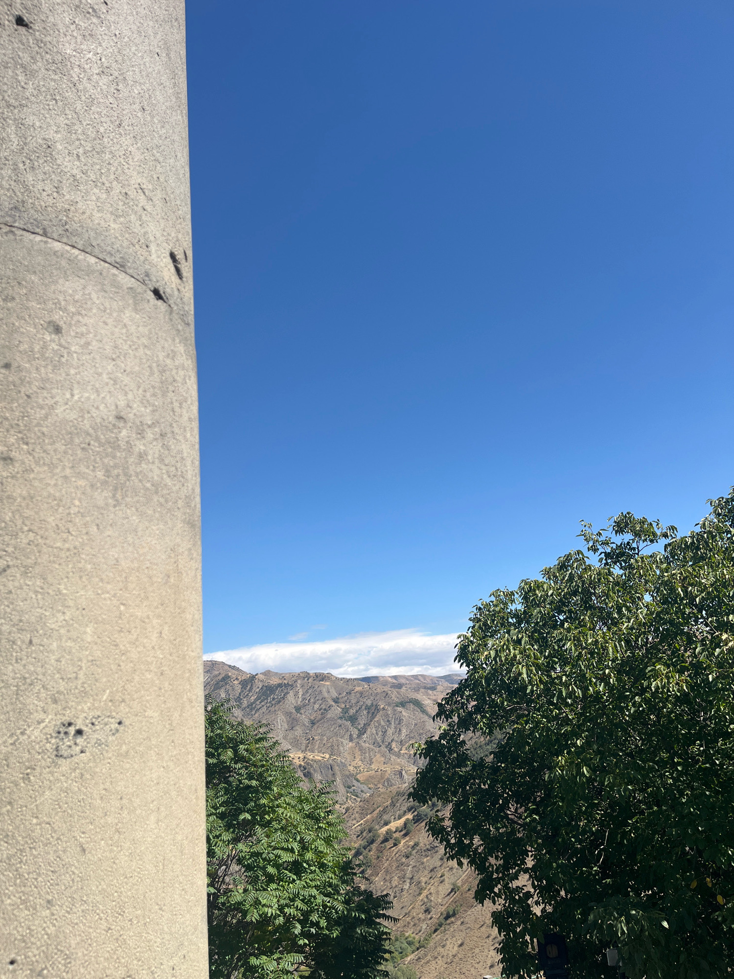 The image, taken from Garni, Armenia, depicts a landscape featuring a stone pillar in the foreground, a lush green tree, and a rugged, mountainous terrain under a clear blue sky. The stone pillar, textured and aged, occupies the left side of the frame, partially obscuring the view of the natural scenery beyond. To the right, a dense canopy of green leaves from a tree branches into the frame, adding a touch of vibrant life to the scene. The background reveals rolling hills and mountains, their slopes etched with shadows and light, suggesting a dry, arid climate. A thin band of white clouds stretches across the horizon above the mountains. The overall atmosphere is one of natural beauty and historical significance, with the ancient stone pillar hinting at the rich past of Garni.