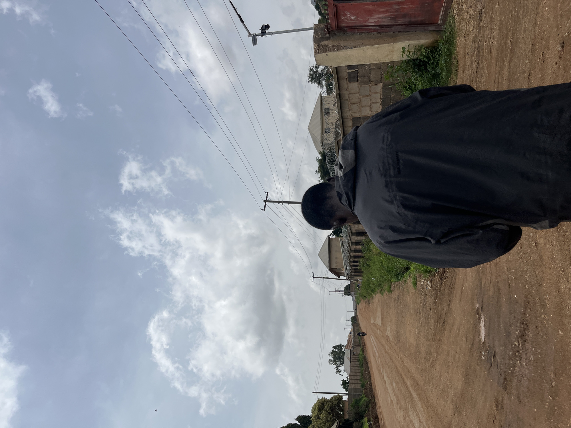 A rear view of a person wearing a dark jacket walks on a dirt road in Bukuru, Nigeria. The road is lined with buildings and vegetation. In the distance, a motorcycle is visible. The sky is overcast with clouds.