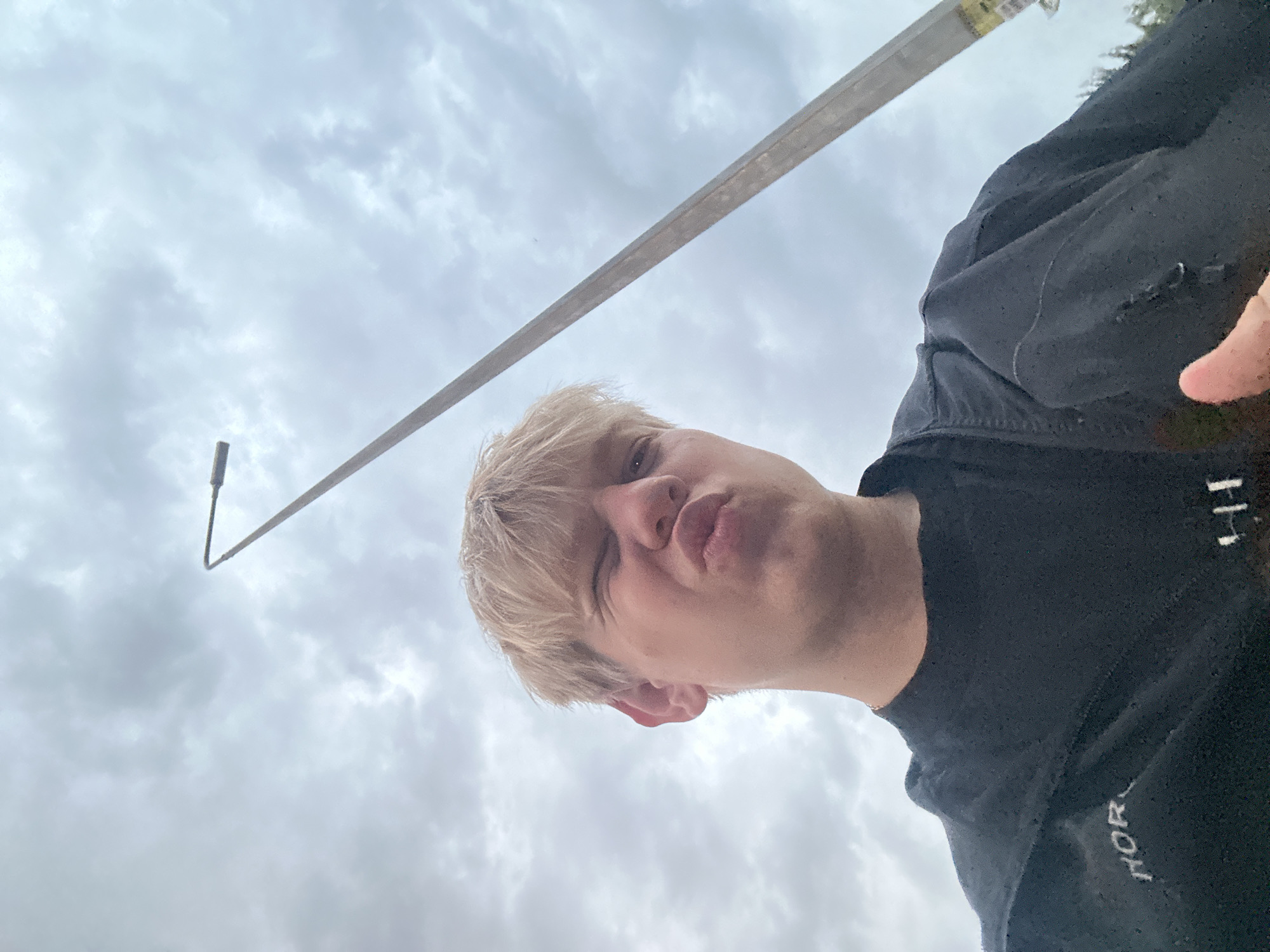 The image shows a young Caucasian male with short, light blonde hair posing for a selfie against a cloudy sky. He is wearing a black t-shirt with some white lettering visible on the chest. The man is making a pouting face with one eye closed. A tall, thin street lamp pole extends diagonally across the frame from the upper right to the lower left. The scene appears to be outdoors, possibly in the city of Khmilnyk, Ukraine, given the context. The angle of the shot is from below, looking up at the subject and the sky.