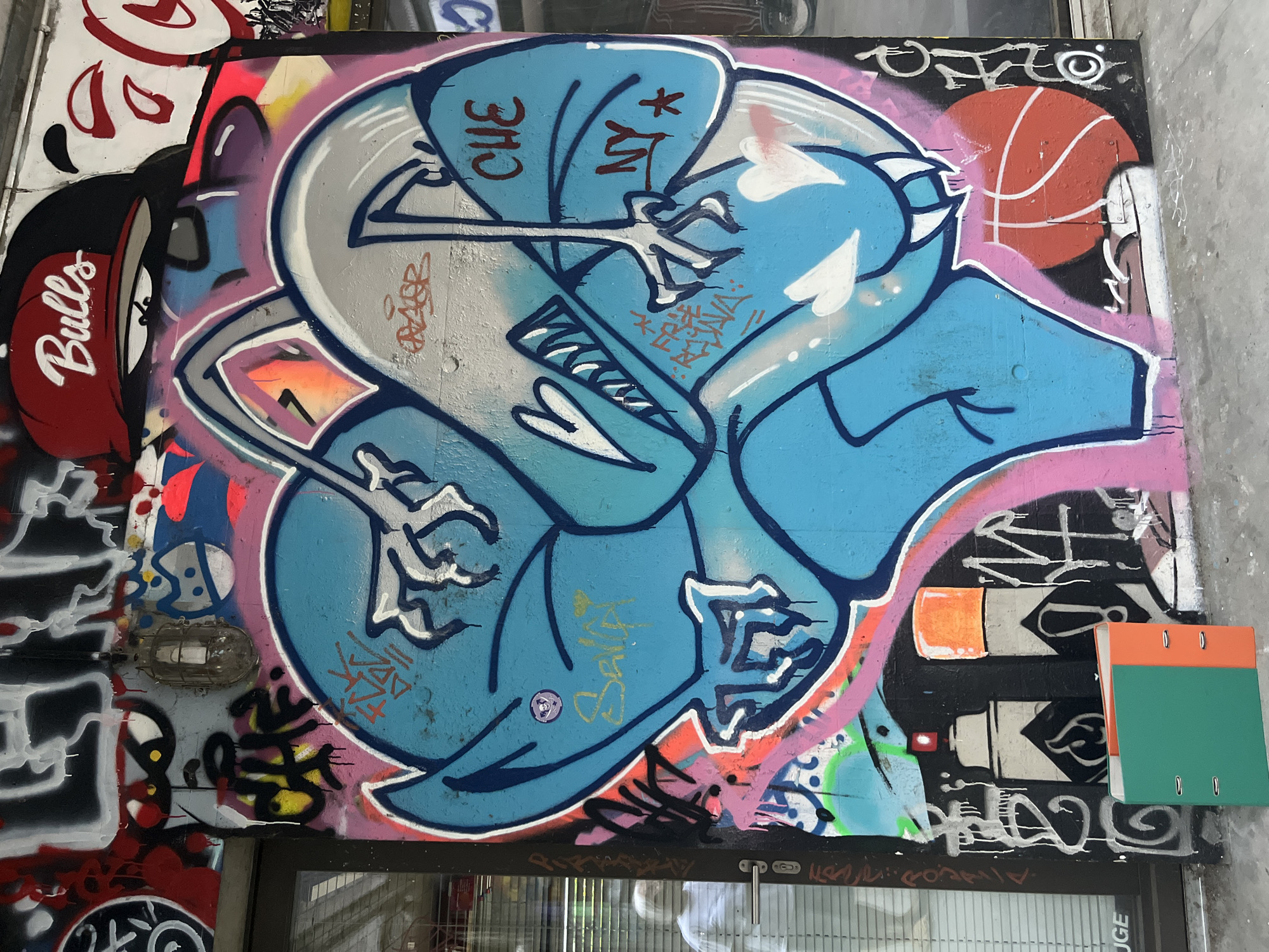 A colorful graffiti mural covers a wall in Geneva, Switzerland. The artwork features a large blue cartoon-like character with sharp teeth and clawed hands. The character is surrounded by various tags and artistic elements, including a red basketball, white hearts, and stylized lettering in shades of pink, orange, and black. Above the main character, a red baseball cap with "Bulls" written in white is visible, along with a cartoon eye. The mural is situated on an exterior wall, partially obscured by a glass storefront on the left, which reflects a person in a white shirt. In the foreground, a green and orange binder folder stands upright on the ground. The overall scene depicts an urban environment with vibrant street art.
