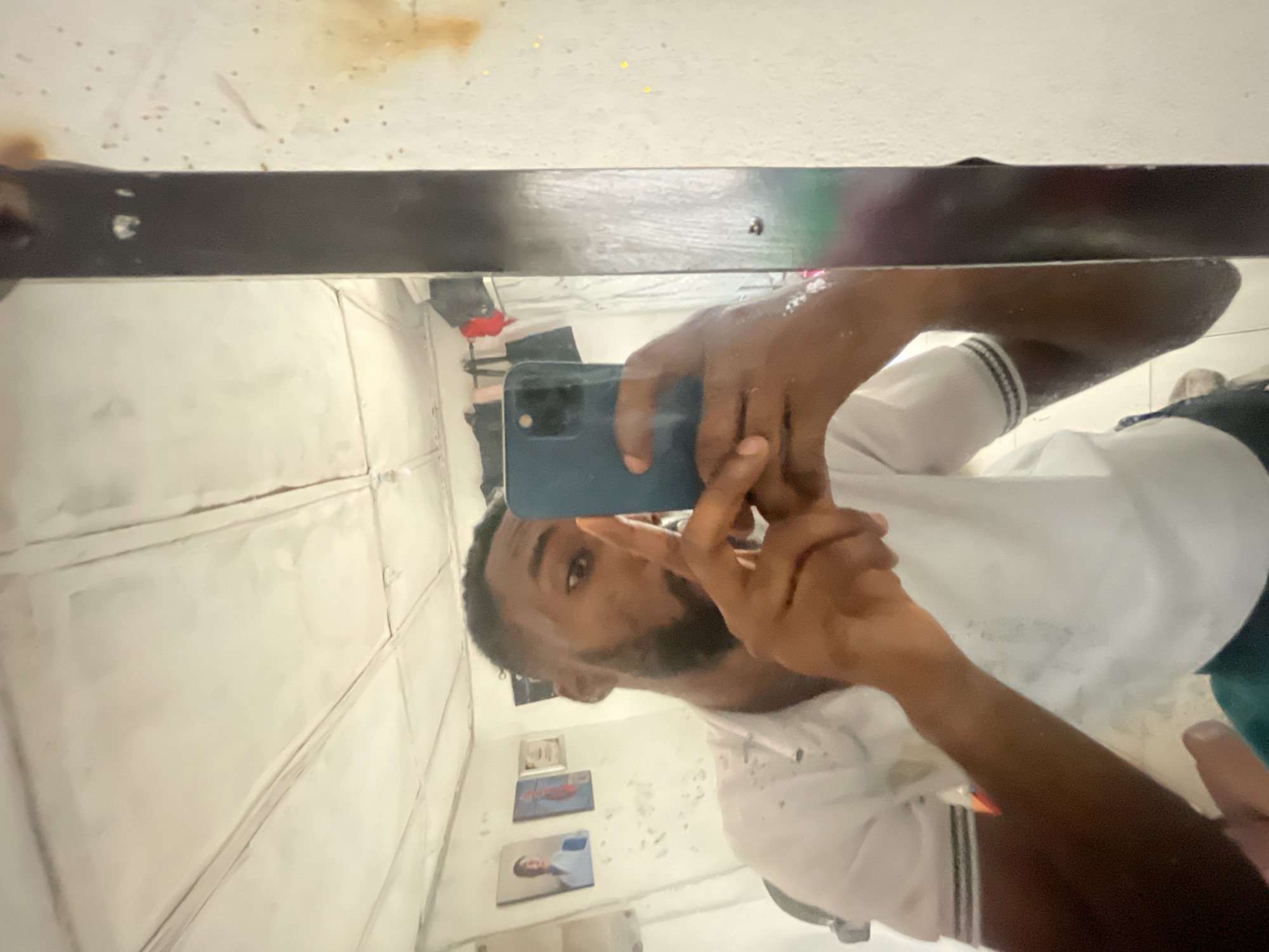 A person takes a photo of themselves in a mirror. The person is dark-skinned, wearing a white short-sleeved shirt, and holding a blue cell phone. The background shows a white tiled wall with framed pictures. The location appears to be an interior space with limited detail visible. There is no indication of the city Lokoja, Nigeria.