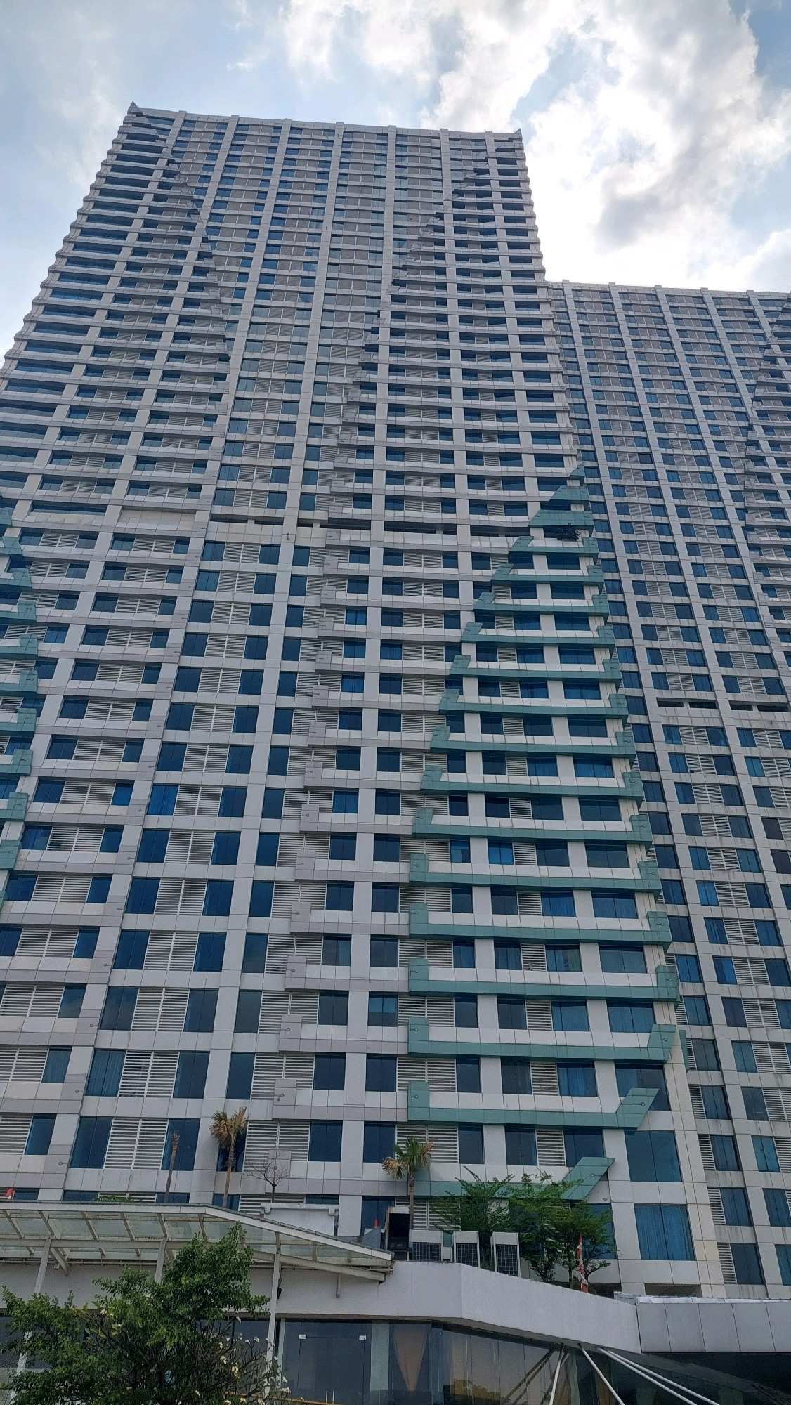 The image shows a low-angle view of a modern, tall building with a facade of alternating light-colored panels and blue windows. The building appears to be a residential or office complex, characterized by its repetitive grid of windows. A distinctive architectural feature is a diagonal, light-green band that runs down the left side of the building, creating a zigzag pattern. The sky above is partly cloudy. In the foreground, at the base of the building, there is a structure with a glass facade and a covered walkway, along with some greenery and palm trees. This building is identified as the Kemang Village Residence in Bekasi, Indonesia.