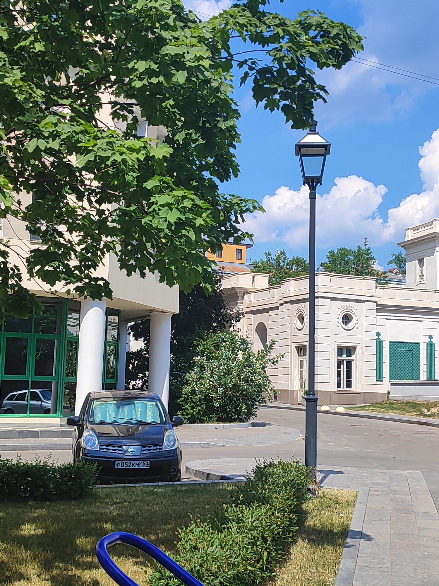 The image depicts a sunny day in a city setting, possibly Moscow, Russia, given the license plate visible on the car. In the foreground, a dark-colored Nissan Note is parked in front of a building with large green-tinted windows and white pillars. The car has a license plate that reads "P052KM 150". A street lamp with a dark, rectangular lantern stands to the right of the car. Behind the car and to the right is a building with light-colored stone facade, featuring arched windows and decorative elements. Lush green trees and bushes are abundant throughout the scene, with one large tree's foliage prominently in the upper left portion of the image. The sky is bright blue with scattered white clouds. A paved walkway runs alongside the street. The overall scene suggests a well-maintained urban environment.
