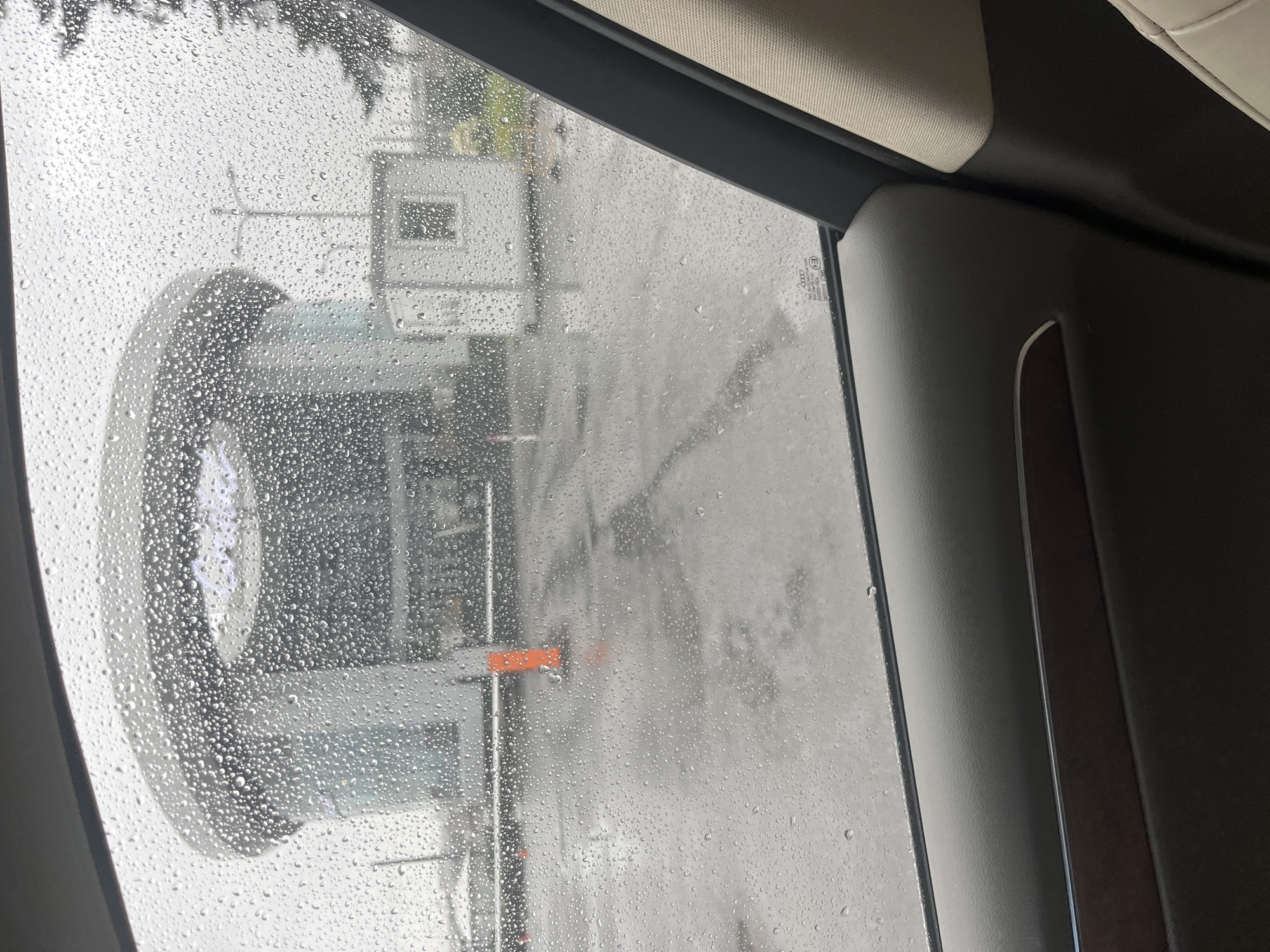 The image shows a view through a rain-streaked car window of an exterior scene. The foreground of the image is dominated by the interior of the car, specifically the car door trim. The middle ground features a wet, paved area with orange traffic cones. In the background, a large, modern, circular building with a glass facade is visible. Adjacent to this building is a small, white, kiosk-like structure. The sky is overcast, indicating a rainy or cloudy day. The overall impression is of a somewhat dreary, urban environment, possibly a parking lot or the grounds of a commercial or public building. The presence of rain on the car window suggests the weather is inclement. The architectural style of the circular building and the overall scene do not provide specific clues to its exact location within Saint Petersburg, Russia.