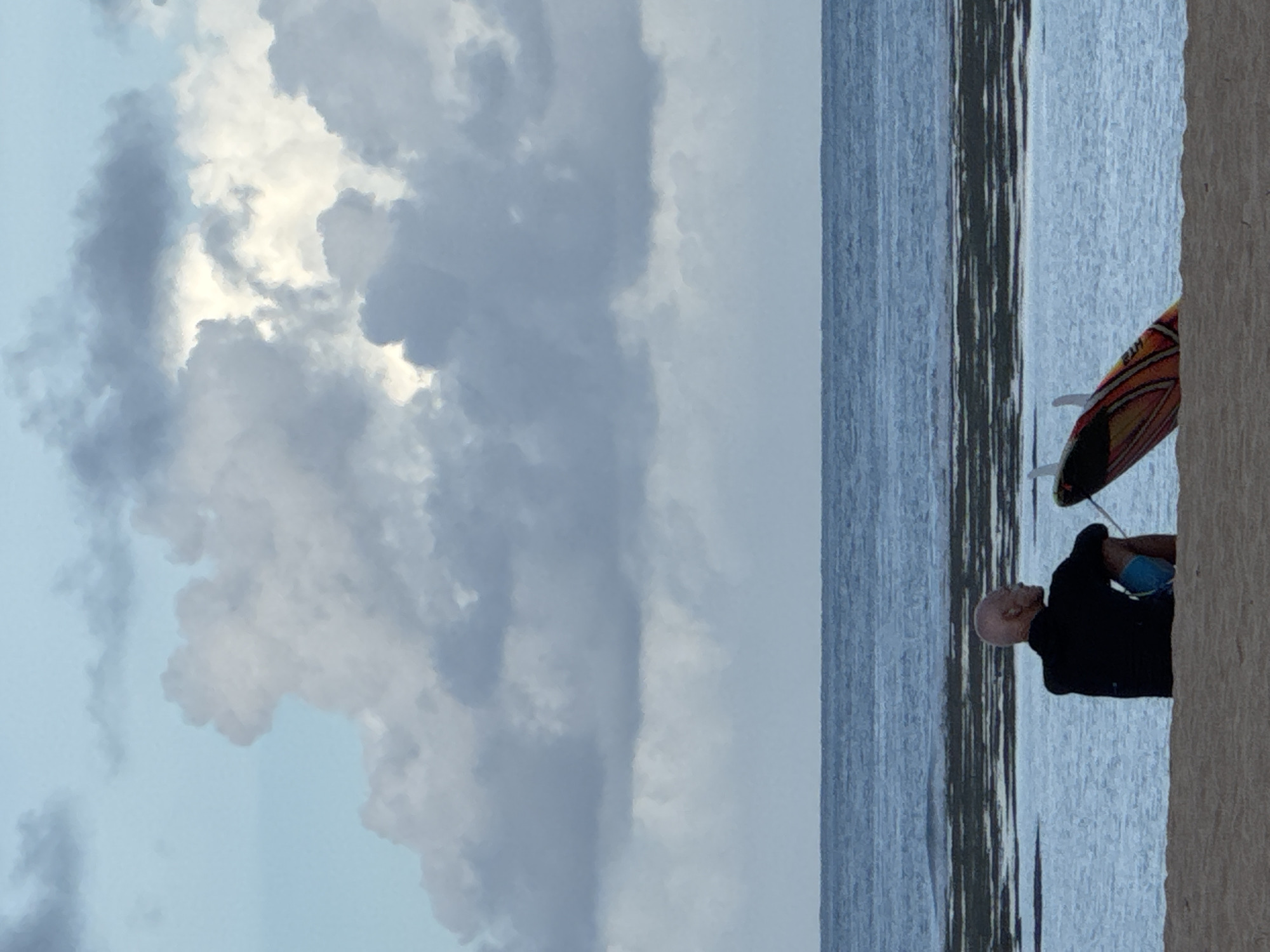 A man stands at the water's edge with a surfboard in Kuta Selatan, Indonesia. The water is calm and the sky is overcast with large clouds. The man is bald and wearing a dark shirt and blue shorts. He is standing next to his surfboard, which is orange and black. The beach is sandy and there is a concrete seawall on the right side of the frame.