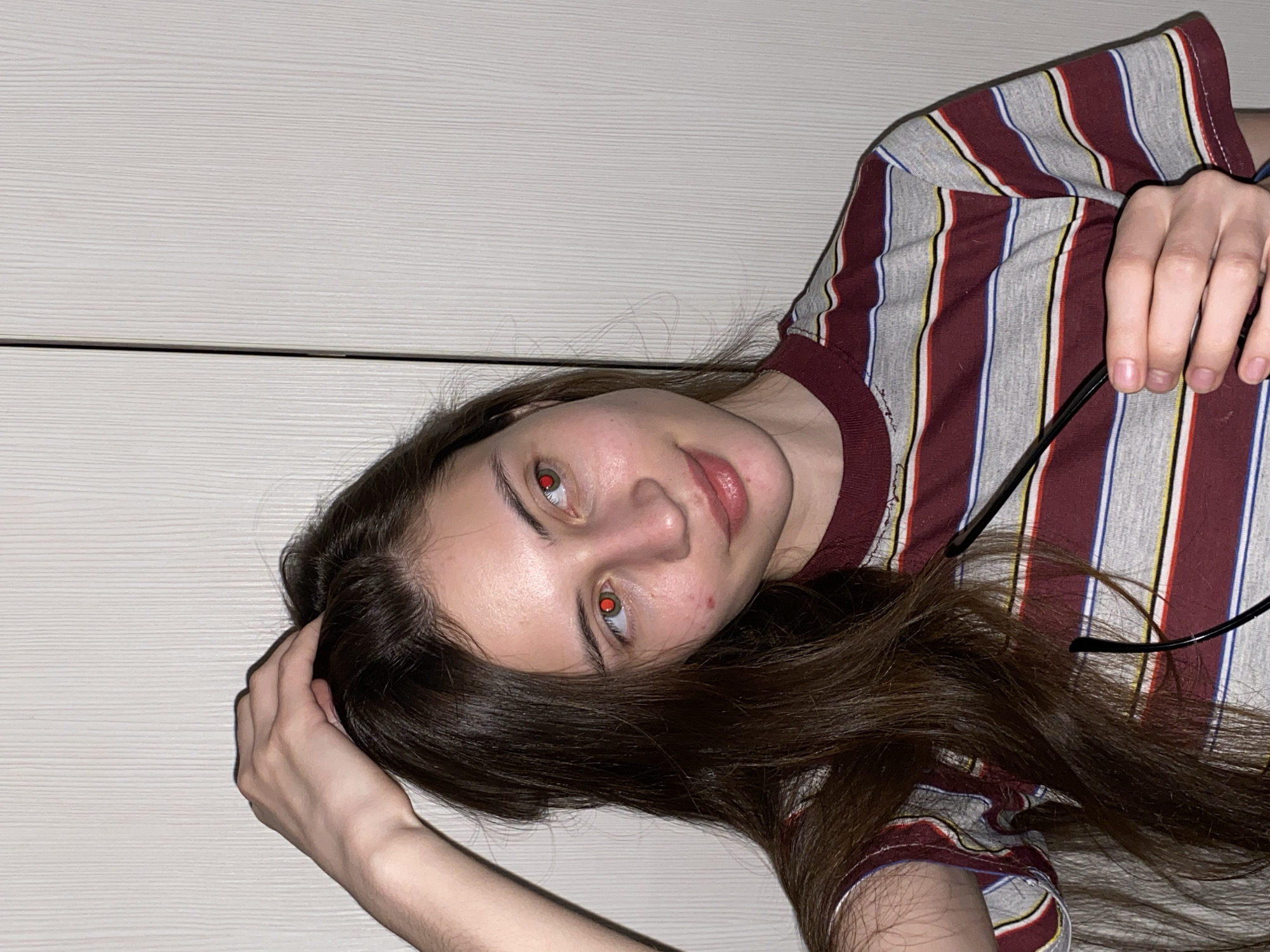 A young person, likely female, is shown in a close-up, rotated portrait, seemingly lying down and looking directly at the camera. She has long, dark brown hair that spreads across a light-colored, horizontally textured background, resembling a wall or headboard. She is wearing a striped t-shirt with a pattern of maroon, gray, white, yellow, and blue vertical stripes.
Her eyes exhibit a distinct red-eye effect, indicating the use of a camera flash, which suggests low-light conditions, likely indoors. She has a subtle, soft expression. One hand is visible near her head, resting on her hair, while the other hand holds a pair of black-framed glasses, which are partially visible against her striped shirt. The setting appears to be a casual indoor environment, consistent with a home in Votkinsk, Russia. There is no visible text in the image.