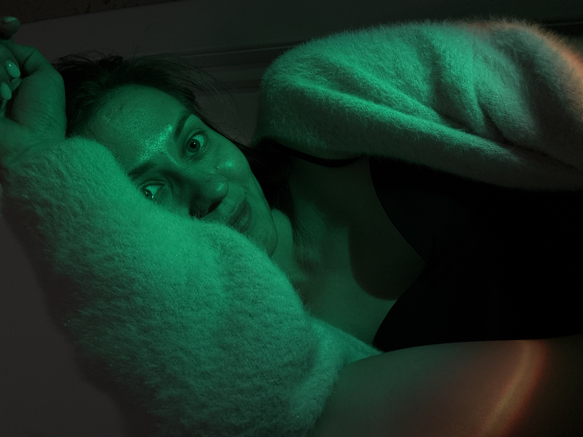 This intimate, low-light portrait features a woman in a reclining position, looking directly at the viewer. Her face, partially framed by dark hair, is illuminated predominantly by a green light source, which creates strong highlights and shadows, giving her skin a distinct emerald hue. Her eyes are wide and engaging, and a subtle smile plays on her lips. Her right hand, adorned with light-colored manicured nails, rests near her head, partially covered by a fluffy, light-colored sweater or blanket. The sweater, which also appears green under the lighting, covers much of her upper body, revealing a dark garment, possibly a top or bra, underneath near her neck and shoulder. In the lower right quadrant of the image, a portion of her leg or thigh is visible, bathed in the same green light but also featuring a soft, distinct reddish-orange glow. The background is dark and indistinct, suggesting an indoor setting, possibly at night or in a dimly lit room. The overall mood is relaxed and direct. No text is visible in the image. The provided context indicates the location as Pervomaysky, Russia.