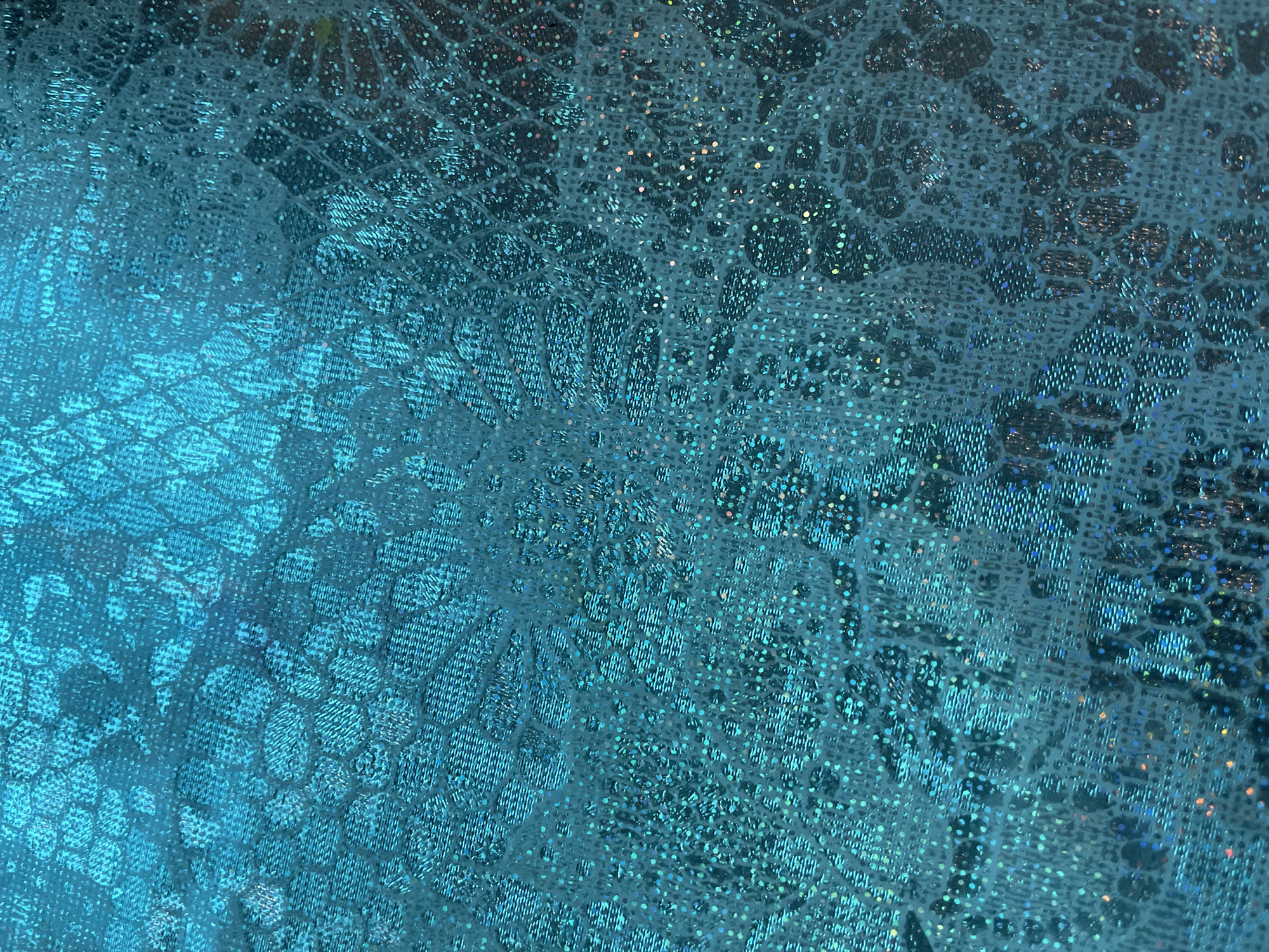 The image shows a close-up view of a blue fabric with a textured pattern that resembles lace or a floral design. The fabric has a slight shimmer, suggesting it might be made of synthetic material or contain metallic threads. The overall tone is cool, with variations in shades of blue highlighting the texture. There is no indication of the location being Olginka, Russia. There are no people, subjects, or objects other than the fabric. There are no actions or interactions. The environment consists solely of the immediate surface of the fabric.