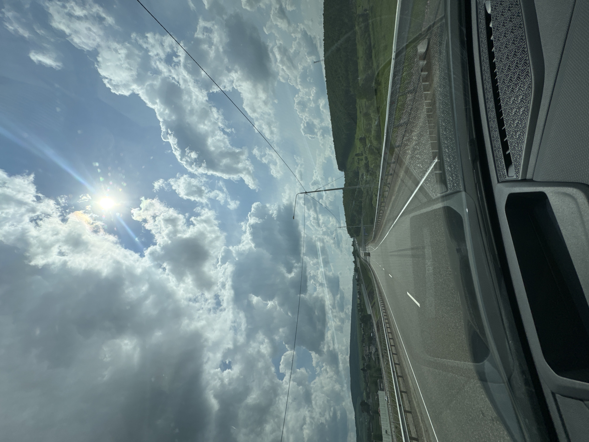 The image depicts a view from the driver's seat of a vehicle traveling on a highway. The road stretches ahead, with clear lane markings and a guardrail bordering the right side. Lush green hills and scattered houses form the landscape in the distance. The sky is dramatic, filled with a mix of dark and light clouds, with the sun shining brightly and creating a lens flare effect. Power lines are visible across the sky. The interior of the car, including the dashboard and windshield, is also captured in the foreground. The scene appears to be a daytime, rural setting in or near the city of Voznesenka, Russia.