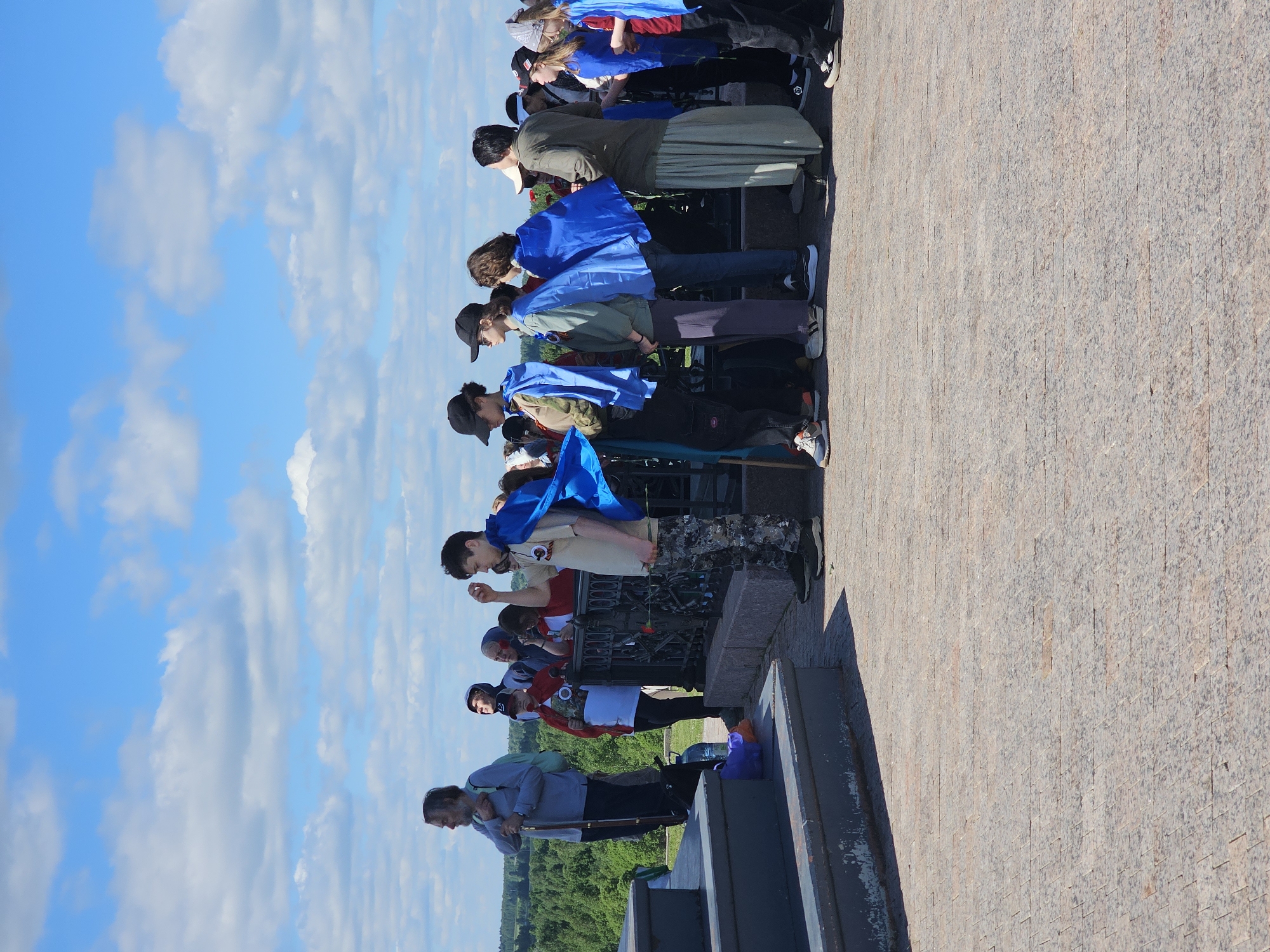 A group of people are lined up along the edge of a stone structure, possibly a memorial or viewing platform. They are near the State Borodino War and History Museum-Preserve in Borodino, Russia. Most of the people are facing forward and appear to be looking at something in the distance. Many wear blue shawls. At least one person is holding a red flower. The background shows a field with trees. The sky is blue with scattered white clouds. The stone structure appears to be paved with small, rectangular blocks. A metal railing is visible along the edge where the people are standing.
