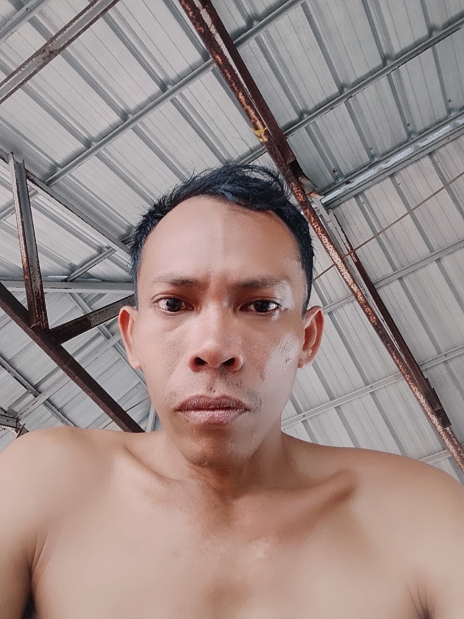 A low-angle, close-up shot of a shirtless man with dark hair and skin. He is staring directly into the camera. The background shows a metal roof structure supported by rusted metal beams. The location is assumed to be in Bukit Ketok, Indonesia, based on external context.