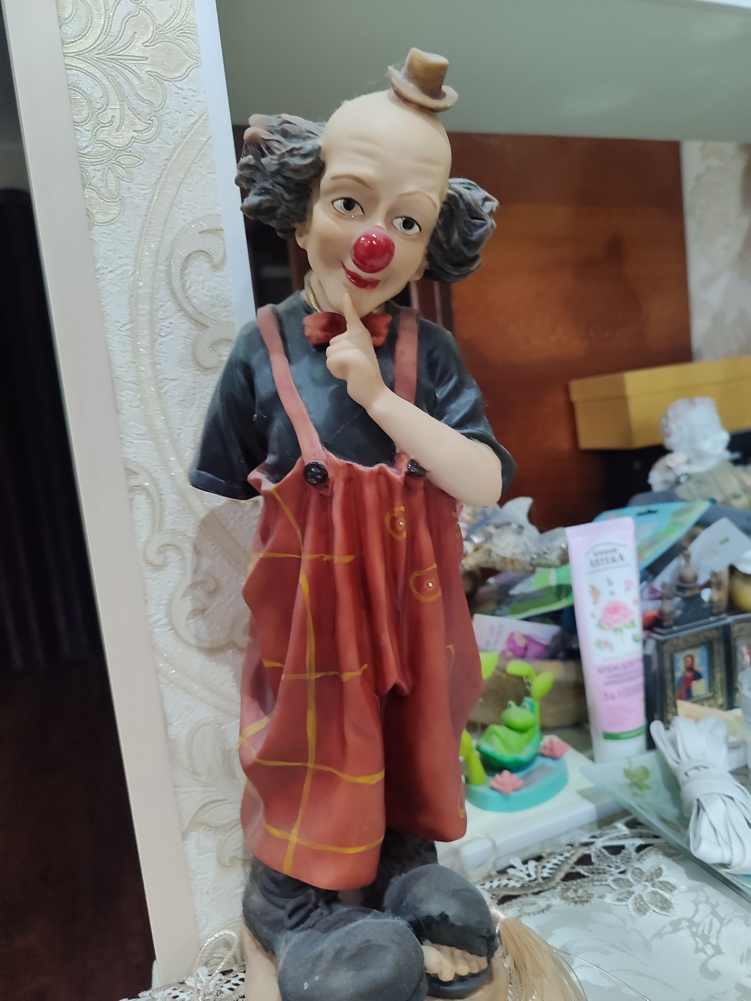 This is a medium shot, eye-level, indoor photograph of a ceramic figurine of a clown. The clown has a bald head with dark curly hair on the sides, a red nose, and is wearing a small brown hat. It has a black shirt with red suspenders and red overalls with yellow plaid patterns. The clown is positioned in the center of the frame, with its right index finger touching its chin, giving it a thoughtful or playful expression.
The background is cluttered, with various objects on a reflective surface, likely a shelf or dresser. To the left of the clown is a cream-colored, textured wall with an ornate pattern. To the right, there are several items including a pink tube of lotion with Cyrillic text and a rose image, a small green frog figurine, a framed picture of a religious figure, and a bundle of white material tied with string. A yellow box is visible in the upper right corner. The lighting suggests it's daytime, possibly indoors with artificial light supplementing natural light. The overall scene appears to be a domestic setting in Tuapse, Russia.