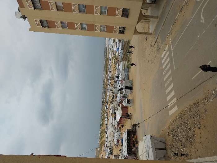 An elevated view captures a street scene bordered by residential buildings and an extensive temporary settlement under an overcast sky in Khan Yunis, Palestinian Territory.
In the foreground, a paved asphalt road features a white pedestrian crossing and a directional arrow painted on its surface. A dark-clad individual stands near the bottom center on the road. A black cable extends diagonally across the scene from the upper left to the lower right. To the left of the road, a sandy slope descends from the edge of a light-colored building, where a white tent structure is visible. A young individual in light clothing walks on this sandy slope.
To the right, a multi-story residential building, light brown with distinctive patterned window frames, rises prominently. A white water tank is visible on its roof. The ground floor of this building is open, supported by columns, and a vehicle is partially visible beneath these arches.
Beyond the road, a sprawling landscape of temporary shelters, tents, and rudimentary structures extends into the distance, predominantly white and grey against the light brown sandy ground. Scattered individuals are present within this area. Two individuals walk past a market-like stall with goods displayed near the left midground. Further right, multiple individuals are seen walking across the sandy terrain amidst the temporary structures. The distant horizon reveals more of the sandy landscape and dense habitation under a grey, cloudy sky.