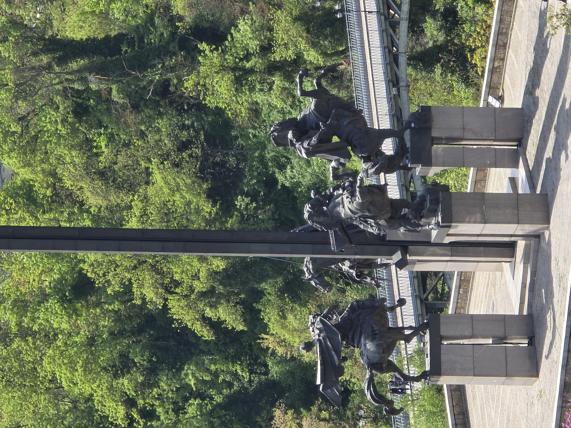 The image depicts a monument featuring bronze sculptures of figures on horseback. The sculptures are mounted on stone plinths or pillars, and they are positioned along a pathway or sidewalk adjacent to a forested area. Above the sculptures, a metal bridge structure is visible. The specific location is not identified as "Unknown, Unknown," but rather as a city for which there is not identifying information provided in the image. The sculptures are the primary subjects, and the background consists of a dense green forest and architectural elements of the bridge and pathway. The day's lighting casts shadows that fall across the pathway.