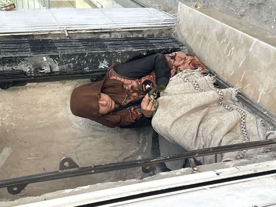 A single individual is seated in an urban alcove in Quetta City Tehsil, Pakistan. The person, appearing young, wears a loose brown head covering, a black long-sleeved shirt, and a brown and beige camouflage-patterned vest with a visible patch on the right chest. A light-colored, checkered patterned blanket or shawl covers their lower body. The individual is actively manipulating a string of green prayer beads with both hands, gazing downwards. The immediate environment includes a weathered, light-colored wall behind the individual. To the right, a dark, corrugated metal shutter is closed and secured with a silver padlock, adjacent to a lighter corrugated metal shutter. A vertical metal bar is present in the left foreground. A light-colored stone or concrete ledge extends horizontally from the right foreground.