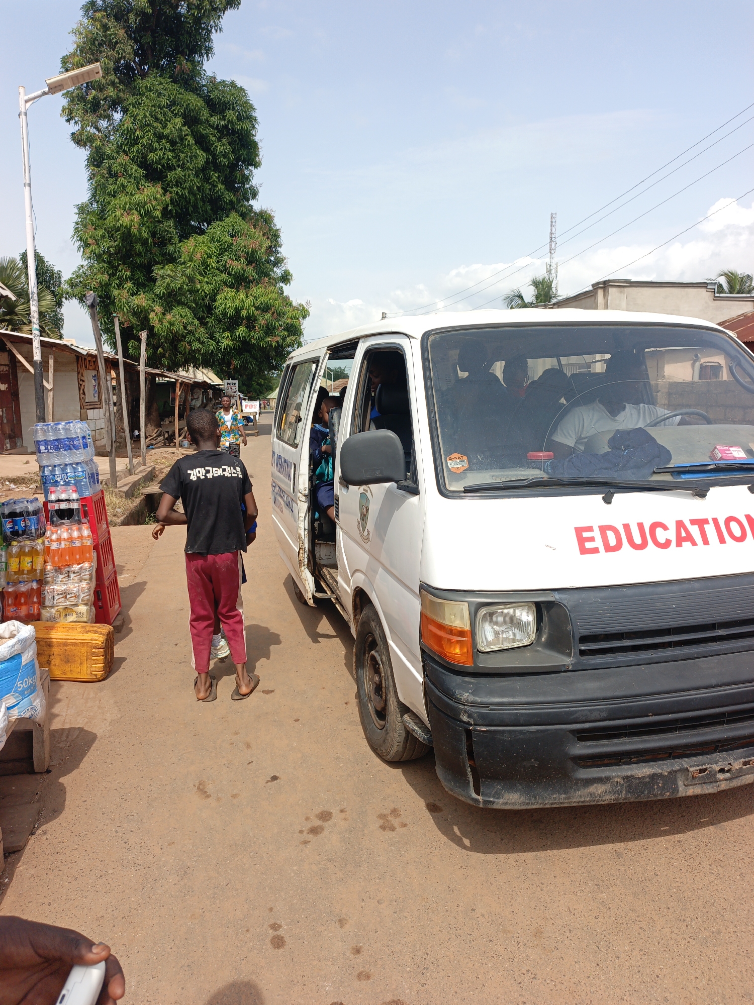 In Akwanga, Nigeria, a white minibus with "EDUCATION" written in red on its front is parked on a dirt road. A young boy in a black t-shirt and red trousers stands facing away from the camera, with his back to the bus, his left arm extended. A partially visible person is behind him. The bus's side door is open, and several children can be seen inside. To the left of the boy, a stall displays stacks of bottled water, sodas, and other beverages, along with yellow and red crates. A large white bag of what appears to be flour is in the foreground. A lamppost with solar panels stands tall behind the stall. The ground is uneven and dusty.