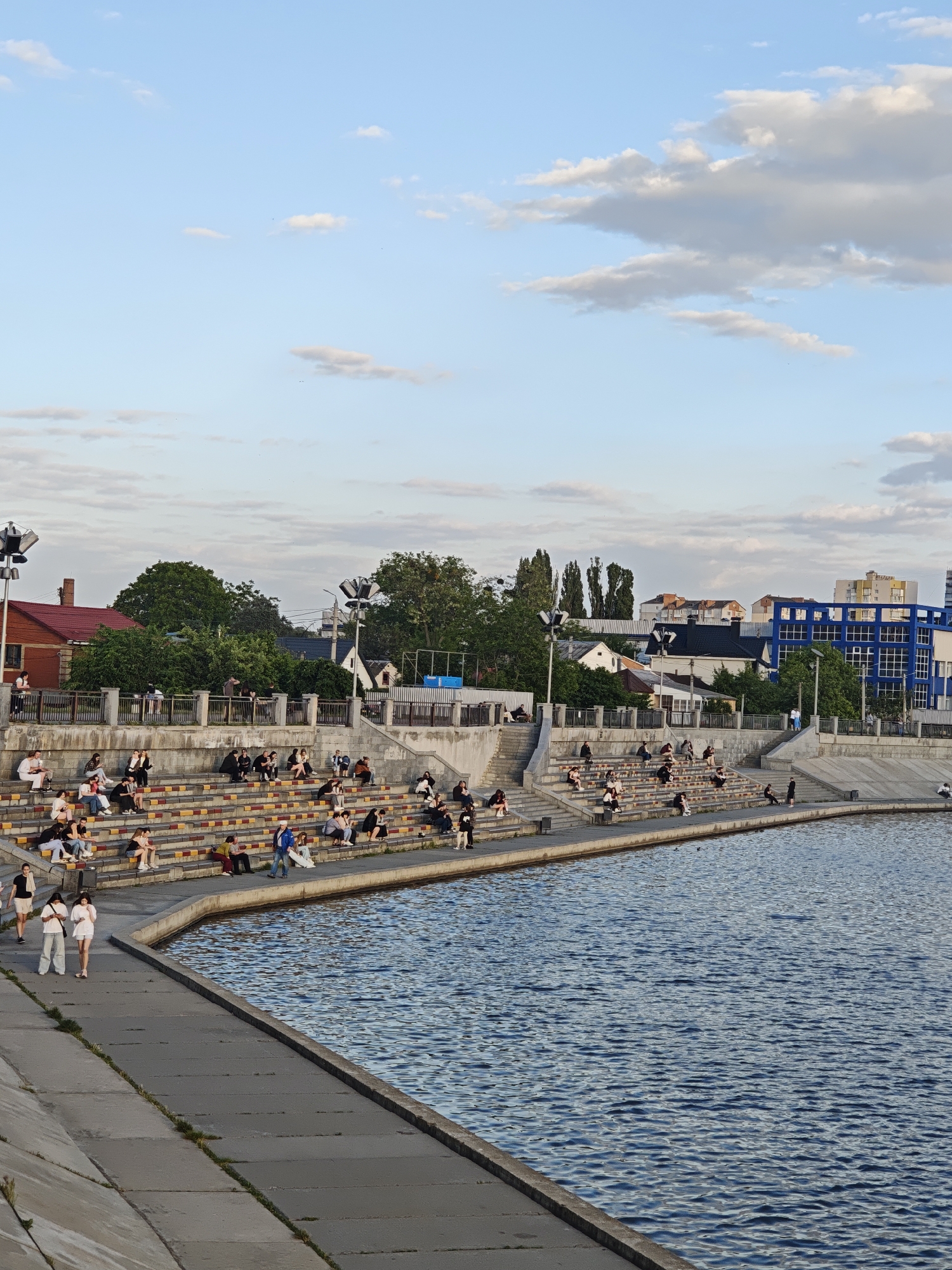 The image captures a scene in Vinnytsia, Ukraine, likely taken at sunset or late afternoon based on the lighting. The foreground is dominated by a body of water, presumably the Southern Bug River, with a concrete embankment along its edge. A small group of two people is strolling along this embankment. Elevated from the river is a tiered structure of steps, serving as an informal amphitheater. These steps are occupied by a diverse group of people sitting and relaxing, mostly in small groups. A mix of clothing styles suggests a varied demographic. Behind the tiered seating, there is a mix of residential and commercial buildings, interspersed with trees. The architecture ranges from older, single-family homes with red roofs to taller, more modern blue buildings. The sky is partly cloudy with patches of blue visible. Streetlights are present along the upper level behind the seating, indicating a public gathering space.