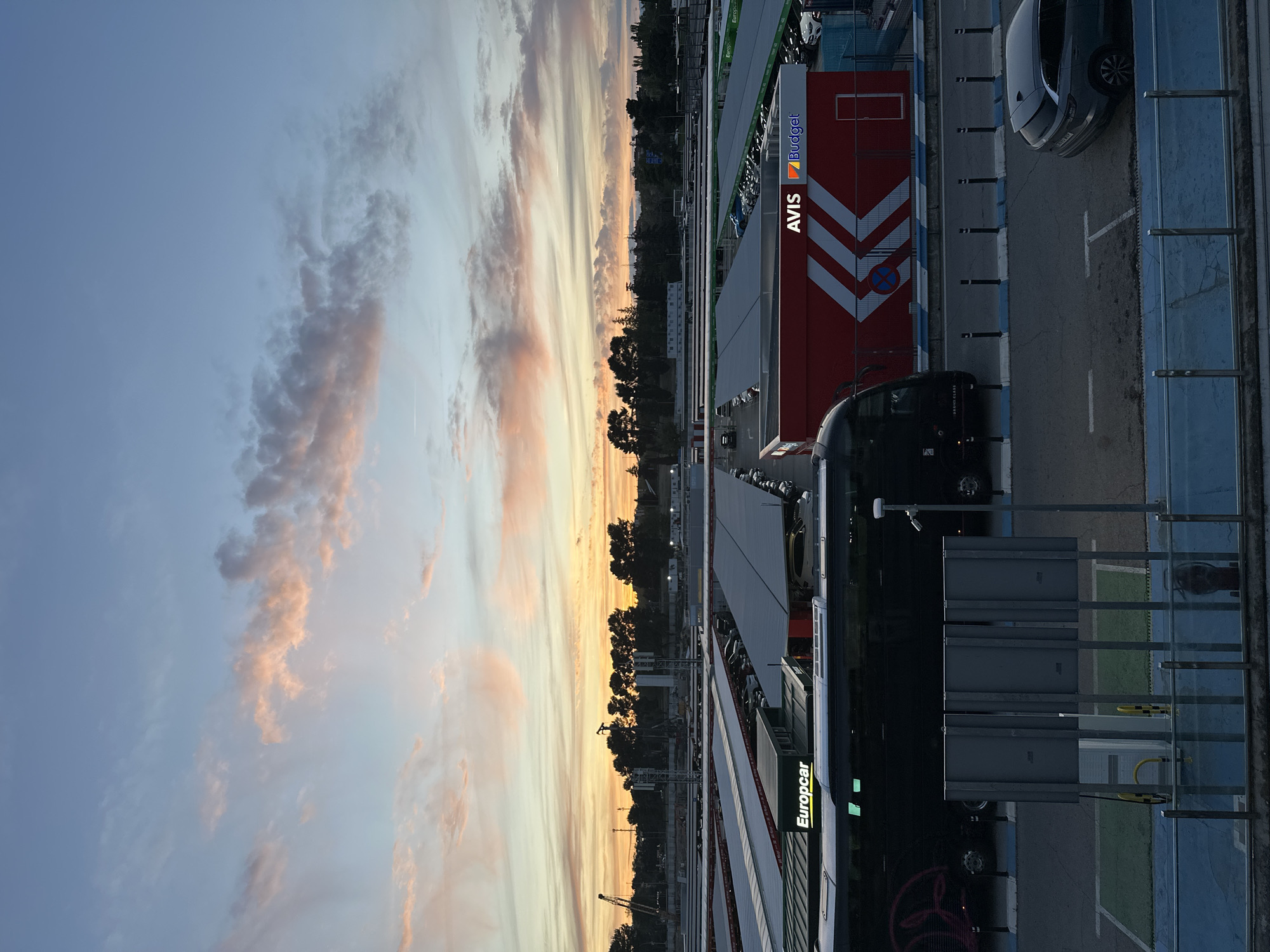The image shows a view of the car rental area at Madrid Airport (MAD) in Spain during sunset. The Avis/Budget rental car facility is visible on the right, and a "Europcar" branded bus is parked on the side, presumably for customer transfers. The backdrop features a colorful sunset with orange and pink hues in the clouds. Several parked cars are visible in the parking lot, and trees and construction cranes are on the other side.