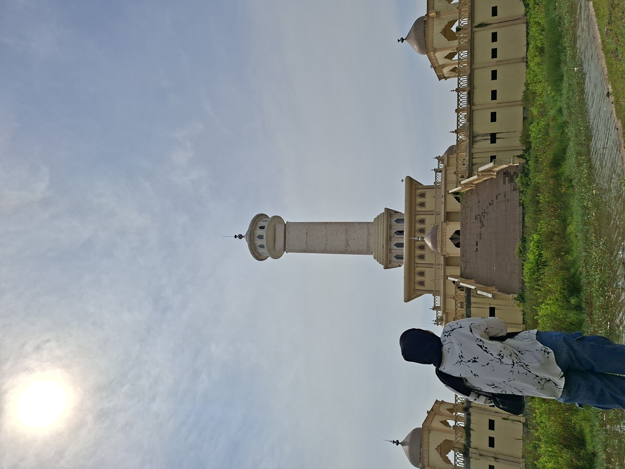 The image captures an outdoor scene featuring a prominent architectural complex and a person, under a bright, partly cloudy sky. The main focal point is a tall, light-colored minaret, slender and multi-tiered, with several ornate balconies and a small dome crowning its top. Adjacent to the minaret, on the right, is a sprawling, multi-storied building, also light beige, characterized by a series of domes, arched windows, and decorative railings, consistent with Islamic architecture.
In the foreground, a person is visible from behind, positioned on the right side of the frame, appearing to gaze towards the architectural structures. They are wearing a dark head covering, a white long-sleeved shirt with black abstract patterns, blue jeans, and carrying a dark shoulder bag.
To the right of the building and the person, a narrow stretch of green grass runs alongside a calm body of water, possibly a canal or moat, which shows faint reflections. The sky occupies a significant portion of the image, displaying a light blue hue with sparse, wispy white clouds. A bright, circular sun is prominent in the upper left section of the sky, indicating a clear, daytime setting. There is no visible text in the image. The provided location context is Keudegeudong, Indonesia.