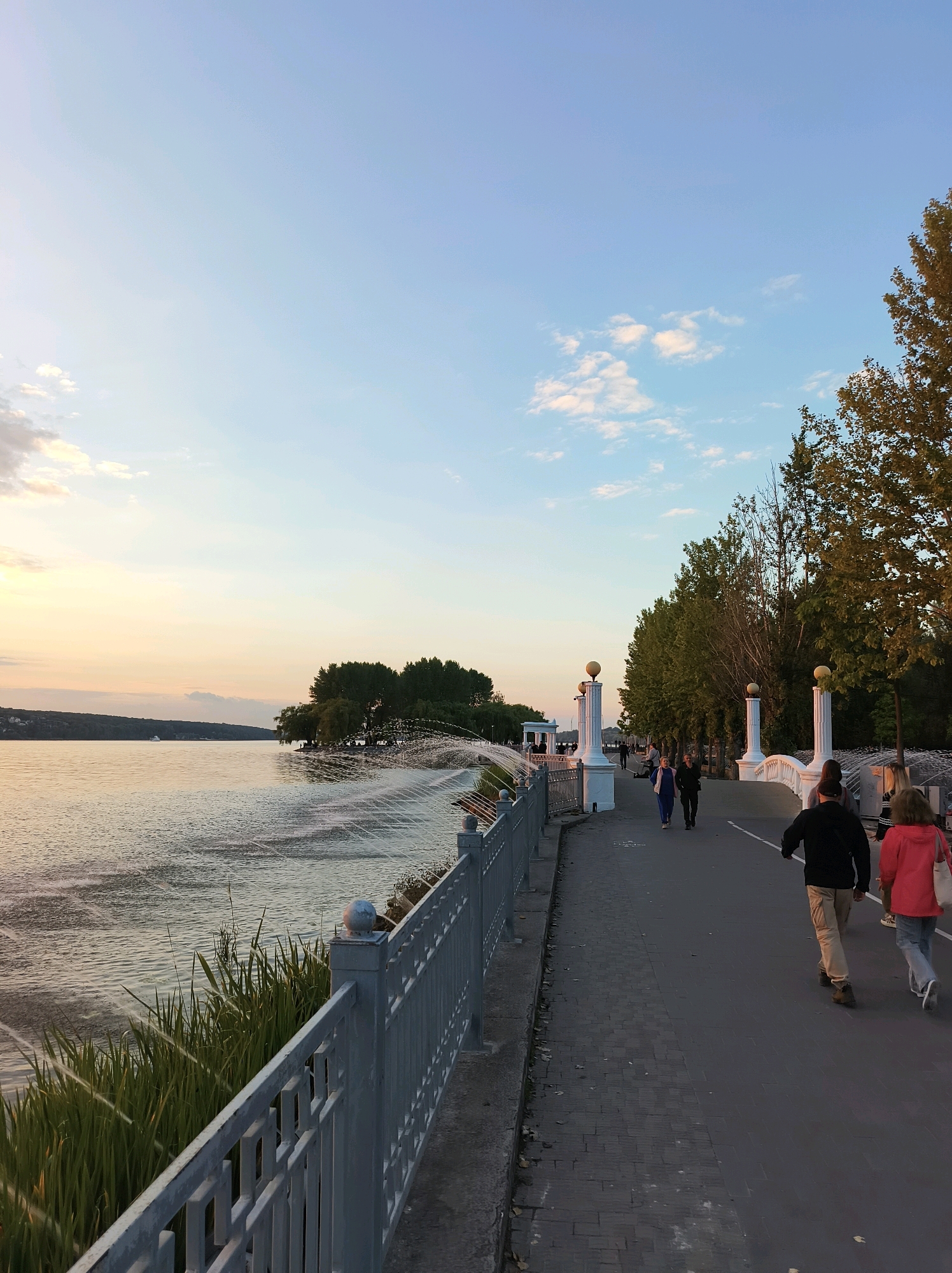 The image depicts a scenic waterfront promenade in Ternopil, Ukraine, during the golden hour. The Ternopil Pond forms the left side of the frame, its surface rippling with a gentle breeze and reflecting the soft hues of the sunset sky. On the right, a paved walkway stretches along the water's edge, lined with trees and ornate white pillars with spherical finials. Several people are strolling along the path, enjoying the evening air. In the foreground, a metal railing separates the walkway from a grassy area and the water, with tall green reeds growing alongside. A series of fountains create a delicate spray across the water in the midground. The sky is a gradient of pale blue and soft orange, with a few scattered clouds. The overall atmosphere is peaceful and idyllic.