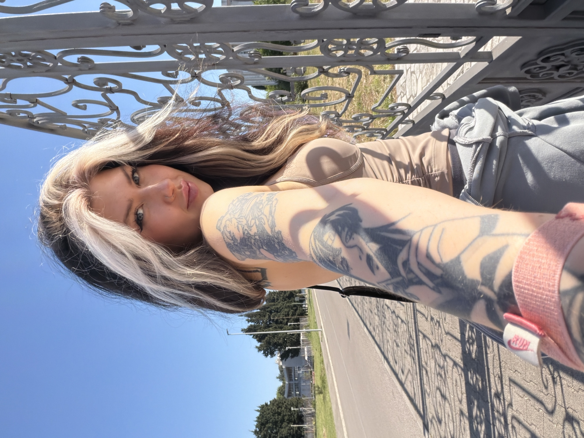A medium shot of a woman taking a selfie. She is fair skinned with dark blonde hair and blonde highlights. She is wearing a tan top and grey bottoms. Her left arm is extended toward the camera and her sleeve is held up by a pink band. There are tattoos on her left arm. She is laying on a grey metal railing. The sky is blue. A road and trees are visible in the background. The location is in Šušanj, Montenegro.