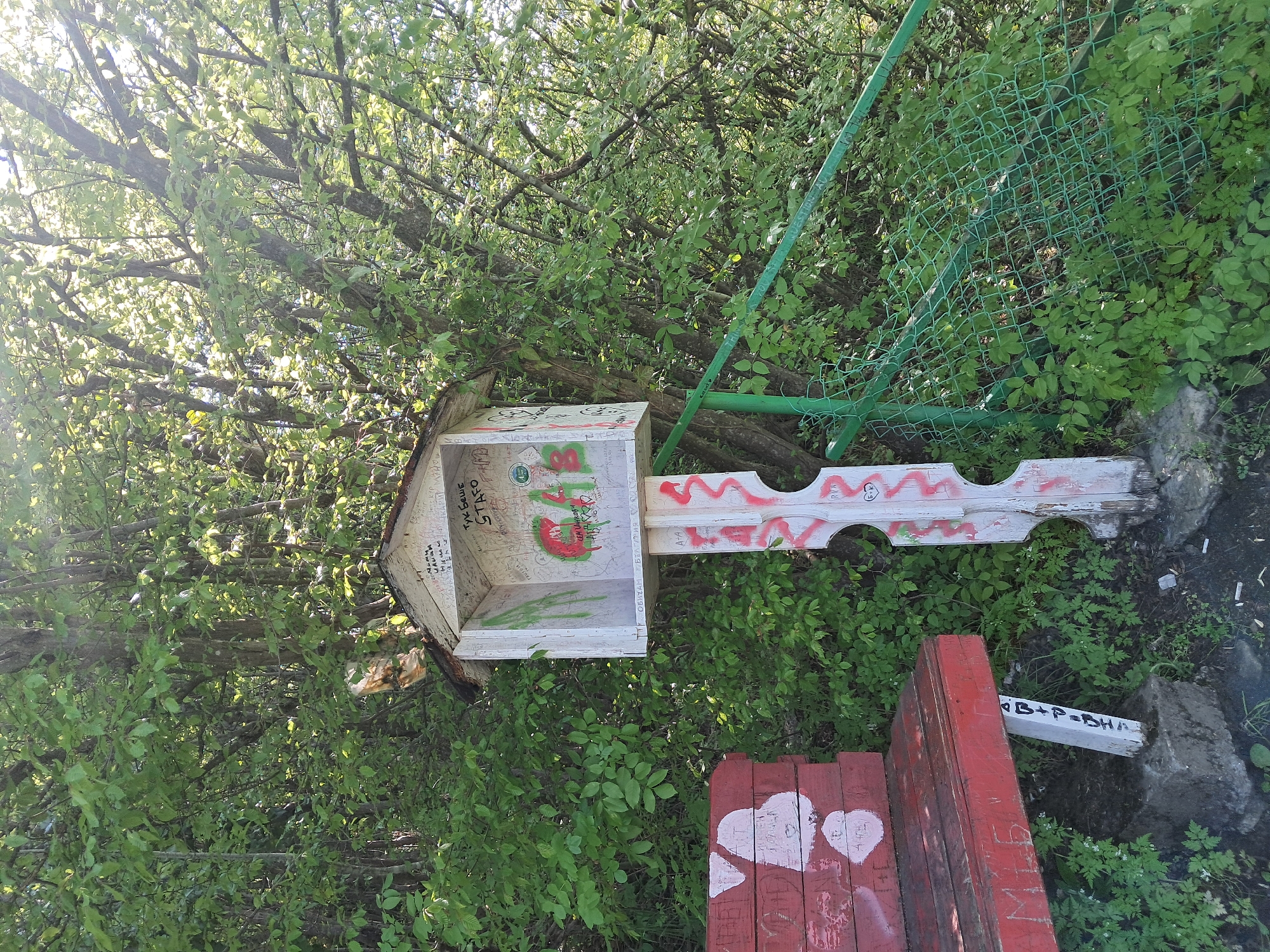 The image presents an outdoor scene, likely in Gesha, Bulgaria, given the Cyrillic script present. The environment is verdant with lush foliage, suggesting a park or wooded area.
In the foreground, there is a structure resembling a birdhouse or a small shrine. It's decorated with graffiti and painted with a red and white theme. The graffiti includes Cyrillic inscriptions and English letters. One reads "Тук Беше STAFO" and another section has text which includes, "ОБИЧАМ БЪЛГАР."
The ground surrounding the structure is covered in green vegetation. A red wooden structure with hearts painted on it is located next to the shrine.
In the background, a green chain-link fence can be partially seen, partially obscured by the foliage. The trees are dense, and their branches are filled with leaves, suggesting it is daytime.
The image has an untended and possibly historical feel, implying that the spot may be a public area where people leave their marks, and that the scene could be a site of remembrance or a local landmark.