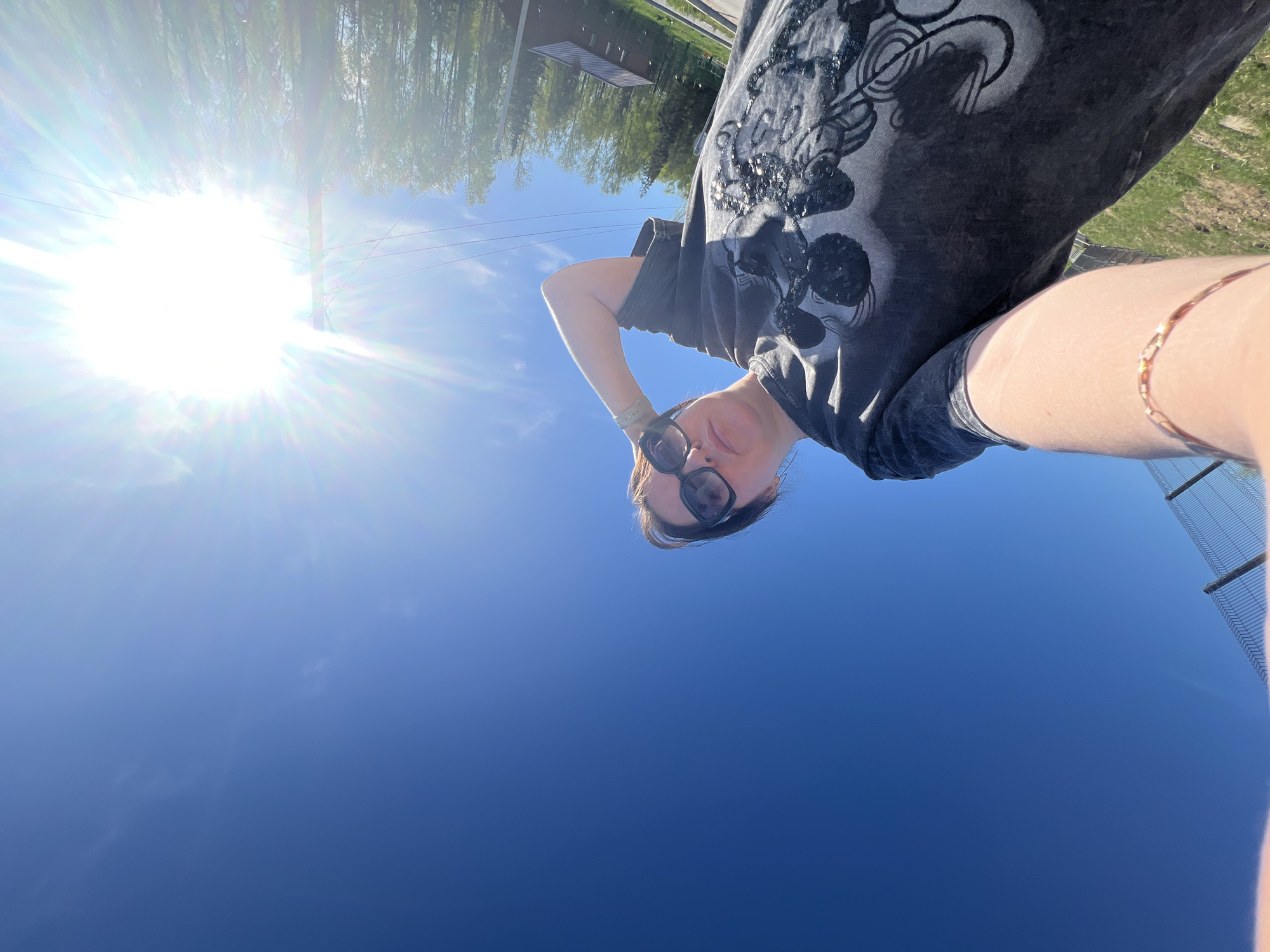 A low-angle selfie shows a person wearing sunglasses and a dark t-shirt with a sequined design. The background is a bright blue sky with the sun visible in the upper left corner, causing lens flare. A line of trees is partially visible at the top. A chain-link fence is faintly visible in the lower right corner. The location cannot be verified.