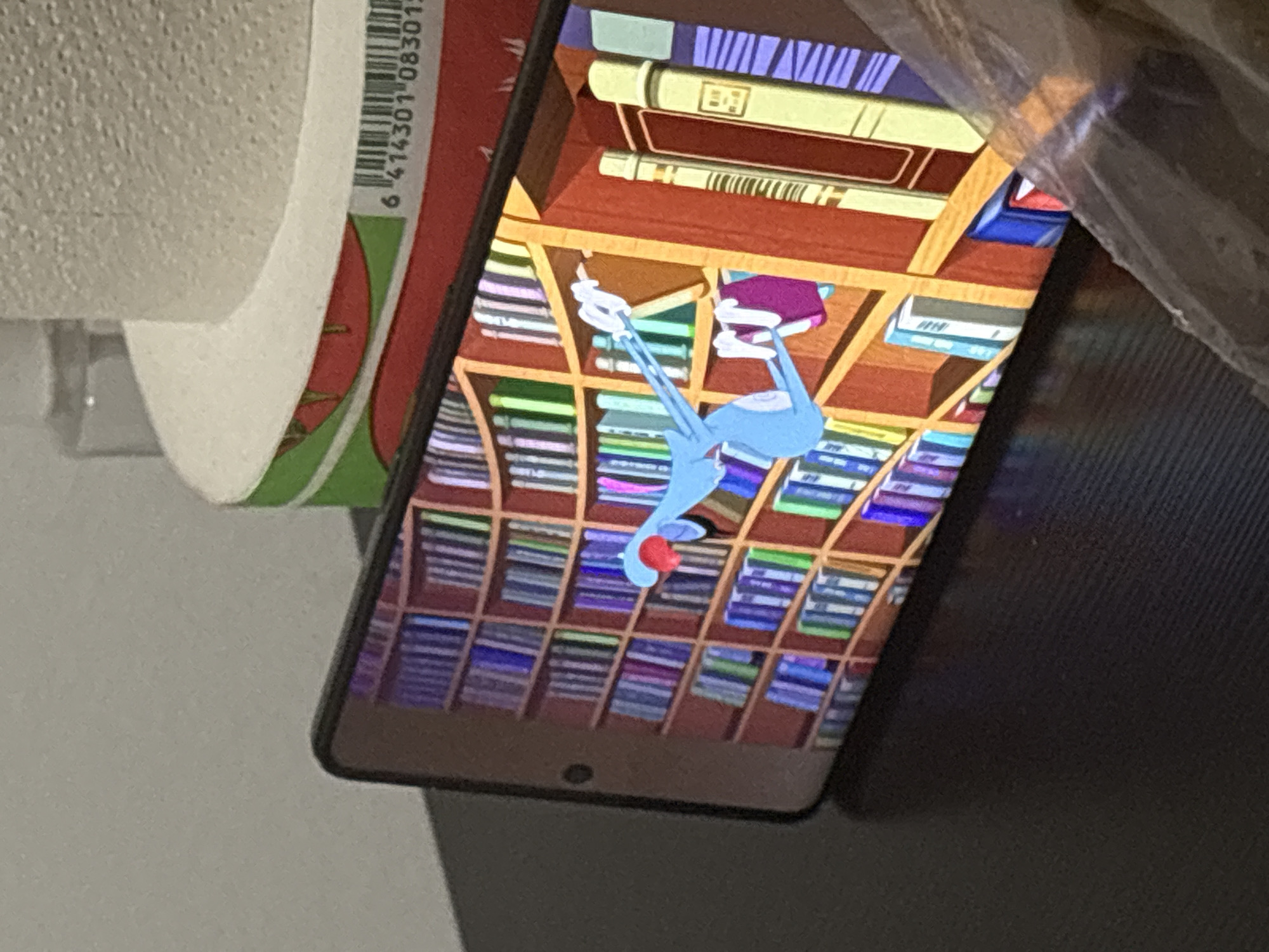 A cellphone is resting on a dark surface, displaying a cartoon on its screen. The cartoon depicts a blue, rabbit-like character with a red nose and outstretched arms, appearing to be reaching for a book on a tall, wooden bookshelf filled with numerous colorful books. The bookshelf extends across the phone's screen. In the background, a portion of a roll of toilet paper is visible, resting on what appears to be a red and green patterned surface. A barcode is printed on this surface. The overall environment is dimly lit, suggesting an indoor setting. The scene is static.