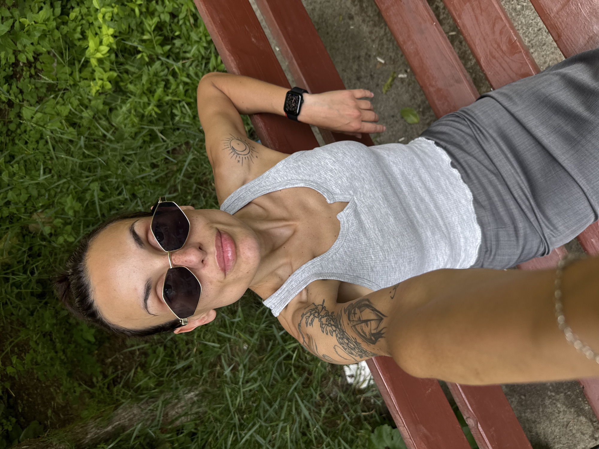A woman is lying on a red wooden bench, seemingly outdoors with grass to the side. She is wearing dark sunglasses, a gray tank top, and gray pants. She has tattoos on both upper arms. On her left wrist, she is wearing a black smartwatch. The photo appears to be a selfie, taken from above. The exact location in Pleven, Bulgaria, cannot be determined from the visual information available.