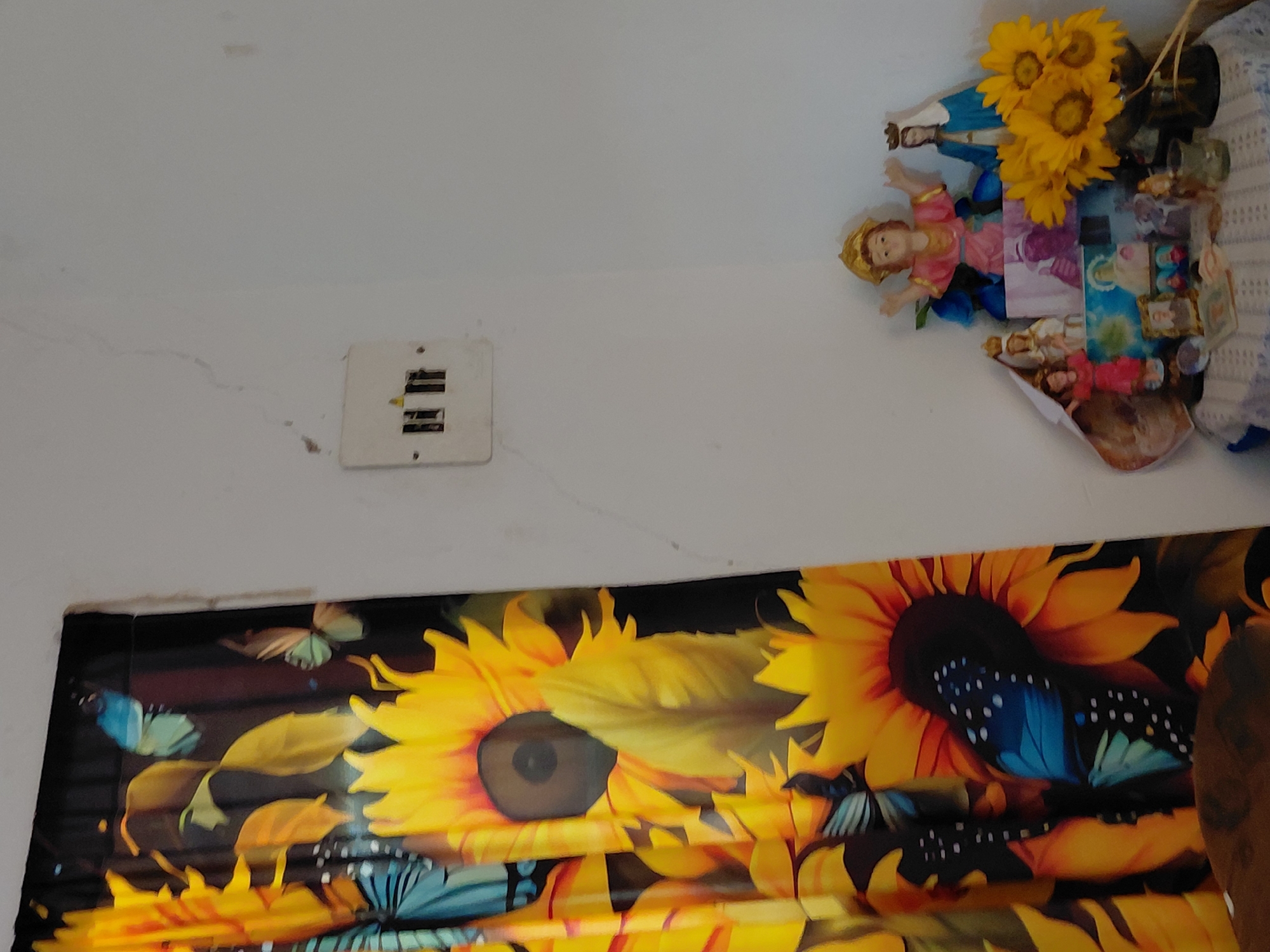 The image depicts a domestic scene, likely a home in Santa Ana, Venezuela. On the left, a curtain with a vibrant sunflower and butterfly print hangs. To the right, on a white wall, is a light switch with exposed wiring. Below the switch and in the corner, a small altar is set up on a table covered with a blue and white checkered cloth. The altar features several religious figurines, including a depiction of the infant Jesus, the Virgin Mary, and other figures. A bouquet of sunflowers is placed to the right of the figurines. The lighting suggests an indoor environment, possibly a living room or bedroom.