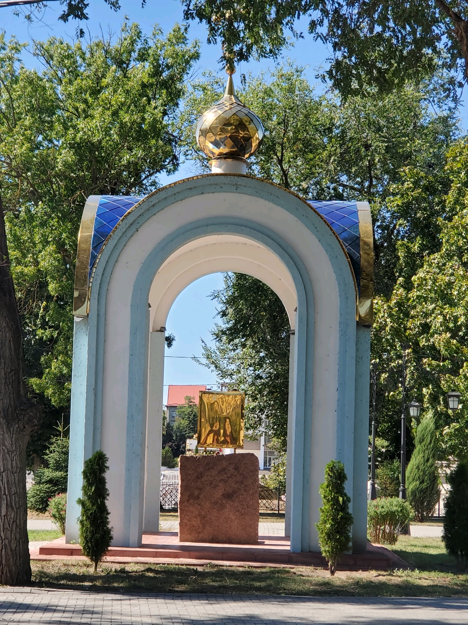 The image depicts an outdoor scene featuring a religious monument. The monument is an arched structure, painted in white and light blue, with a golden dome topped with a cross. Beneath the arch, a golden icon or religious panel is displayed on a reddish-brown stone pedestal. Two small, conical evergreen trees flank the pedestal. In the background, there are trees, a building with a red roof, and a street lamp. The ground in the foreground is paved with cobblestones. The sky is clear and blue, suggesting a sunny day. The location is identified as the Chapel of the Ascension of the Lord in Tiraspol, Moldova, situated near the National Bank of Pridnestrovie.
