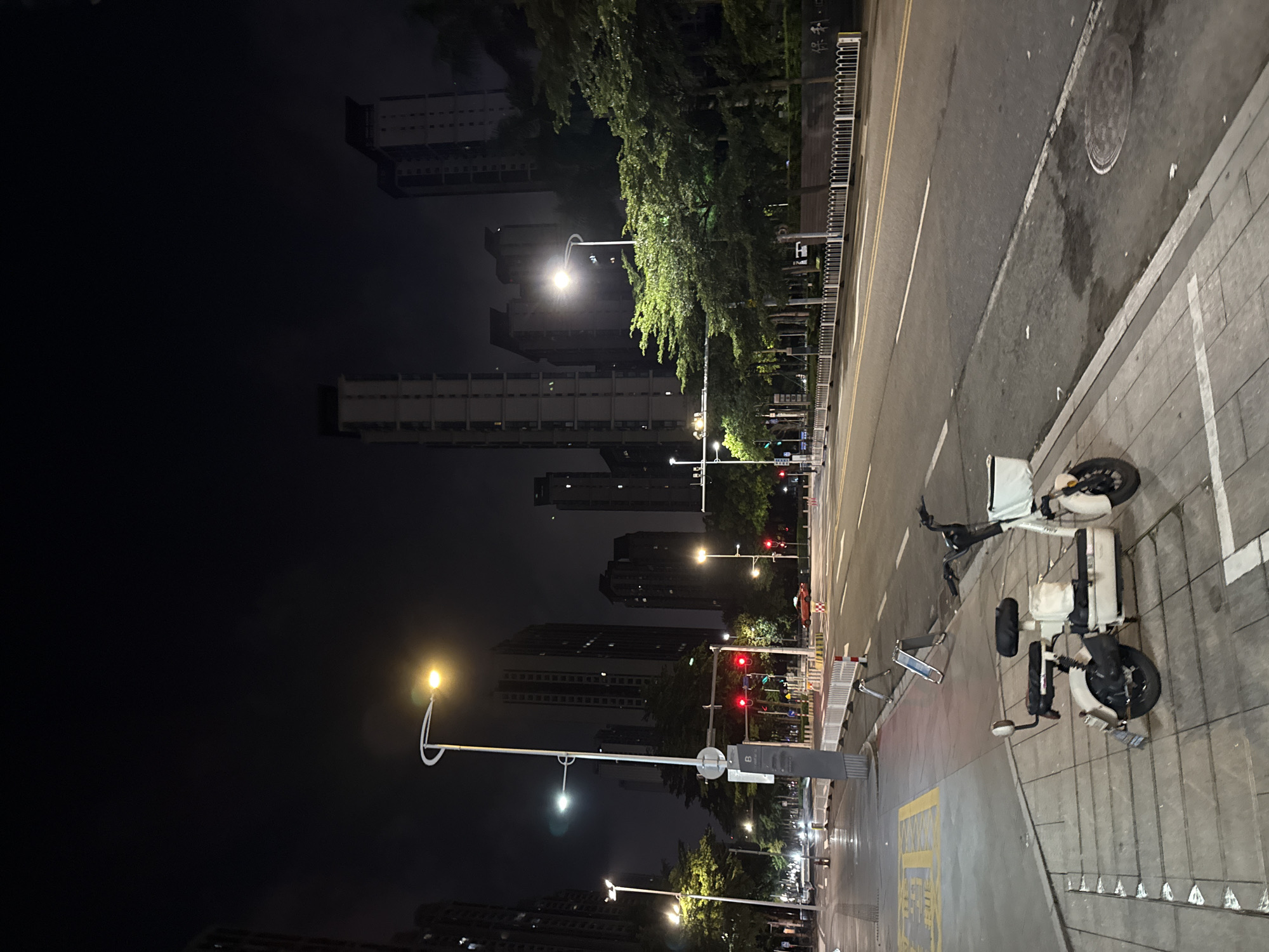 The image shows a nighttime street scene in Haizhu District, China. The perspective is from a low angle, looking up at tall buildings that dominate the dark sky. The buildings are illuminated by streetlights and lights from within the structures, creating a stark contrast with the deep blackness of the night.
A street with yellow lane markings runs alongside a sidewalk. On the sidewalk, a white electric scooter is parked. The scooter appears to be a shared mobility device, common in urban areas. Further down the street, more streetlights illuminate the scene, casting a warm glow. Traffic lights are visible, some showing red, indicating a pause in the flow of vehicles. Trees line the street, their foliage catching the light and appearing lush.
The overall atmosphere is urban and nocturnal, with a sense of quiet activity. The lighting suggests that it is nighttime, and there are no clear indicators of weather conditions beyond the artificial illumination. The presence of apartment buildings and street infrastructure points to a residential or mixed-use urban environment. There is some visible text on a sign near the scooter, but it is too blurry to be legible.