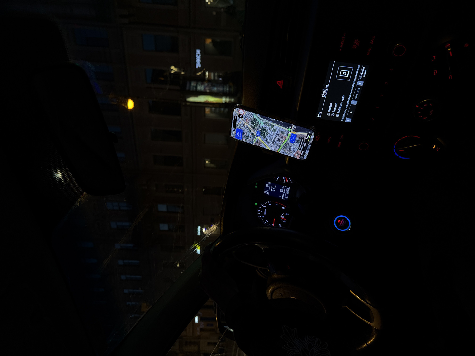 An interior night shot from the driver's perspective inside a car parked on a city street, likely in Saint Petersburg, Russia. The dashboard is illuminated by the car's instruments and the screen of a smartphone mounted on the dashboard, displaying a navigation app. The exterior view through the windshield shows a dimly lit street with buildings lining the opposite side of the road. A street lamp with a yellow glow is visible, casting light on the building facades. The car's engine start button is illuminated red and blue. The car's infotainment screen displays the time as 12:56 AM and the text "NYUSHA", "больно", and "Выбрать чудо" (Choose a miracle), indicating it is playing music or an audiobook. The background buildings have a classic architectural style, suggestive of older European cities, consistent with Saint Petersburg. There are no people visible in the image.