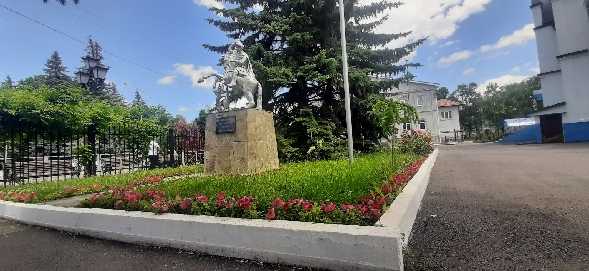 The image shows a public square with a monument, trees, flower beds, and buildings in Yessentukskaya, Russia. In the center is a silver equestrian statue on a stone pedestal. The statue depicts a warrior in armor with a helmet and spear. In front of the monument, there is a flower bed with red and pink flowers, and lush green grass. Behind the monument are tall evergreen trees, and a black metal fence encloses a grassy area with benches. To the right of the monument, there is a gray building with white trim and windows, and a blue and white annex with an entrance ramp. A person in a white shirt is visible near the fence on the left side of the image. The sky is blue with scattered white clouds. The ground in the foreground is asphalt.