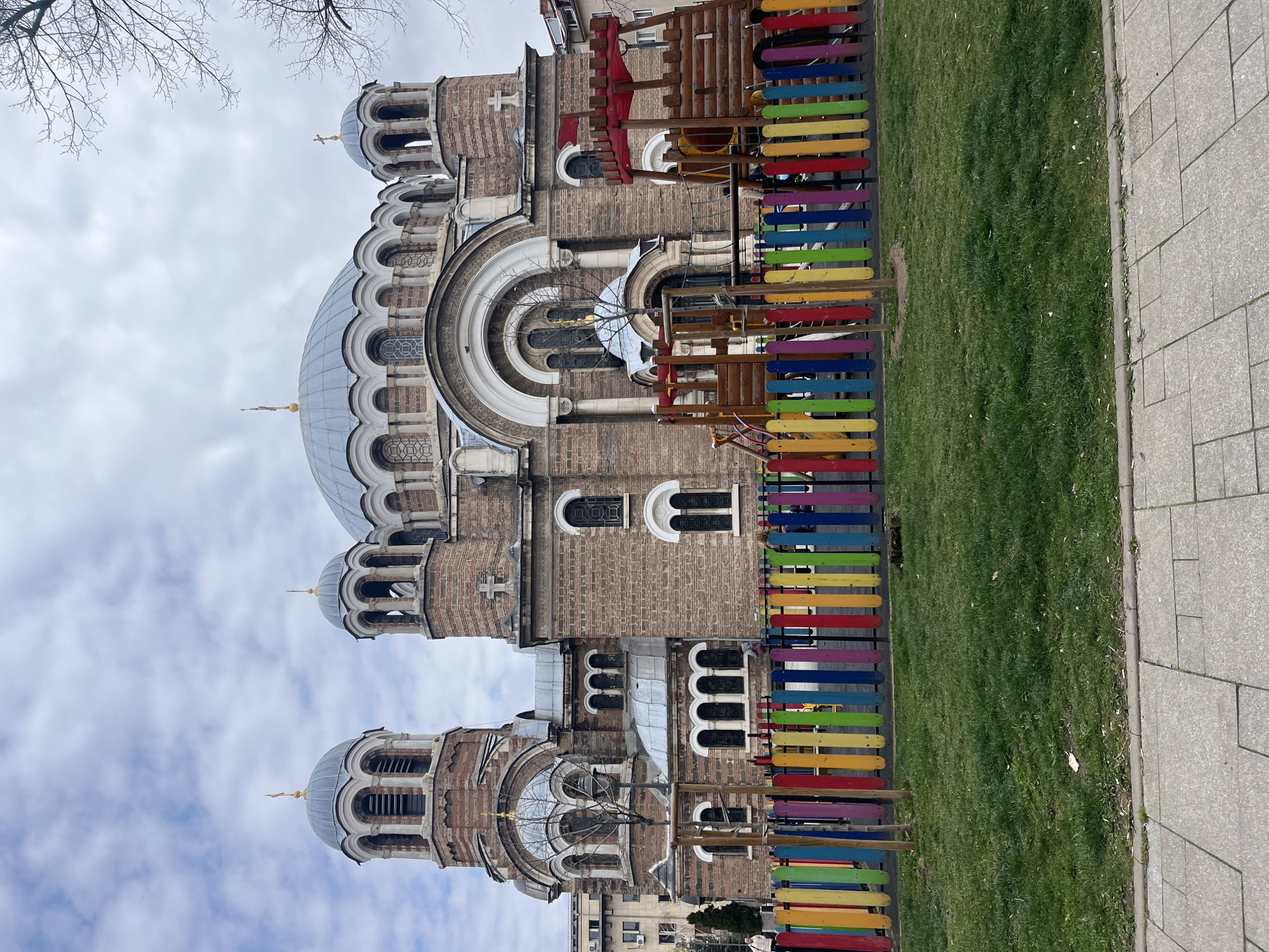 The image shows the Alexander Nevsky Cathedral in Sofia, Bulgaria. The cathedral's dome and several of its smaller domes are visible against a cloudy sky. To the right of the cathedral, there is a playground structure and a colorful fence. Beyond the fence, there is green grass and a paved walkway. A multi-story building is visible in the background. There are no people visible in the image.