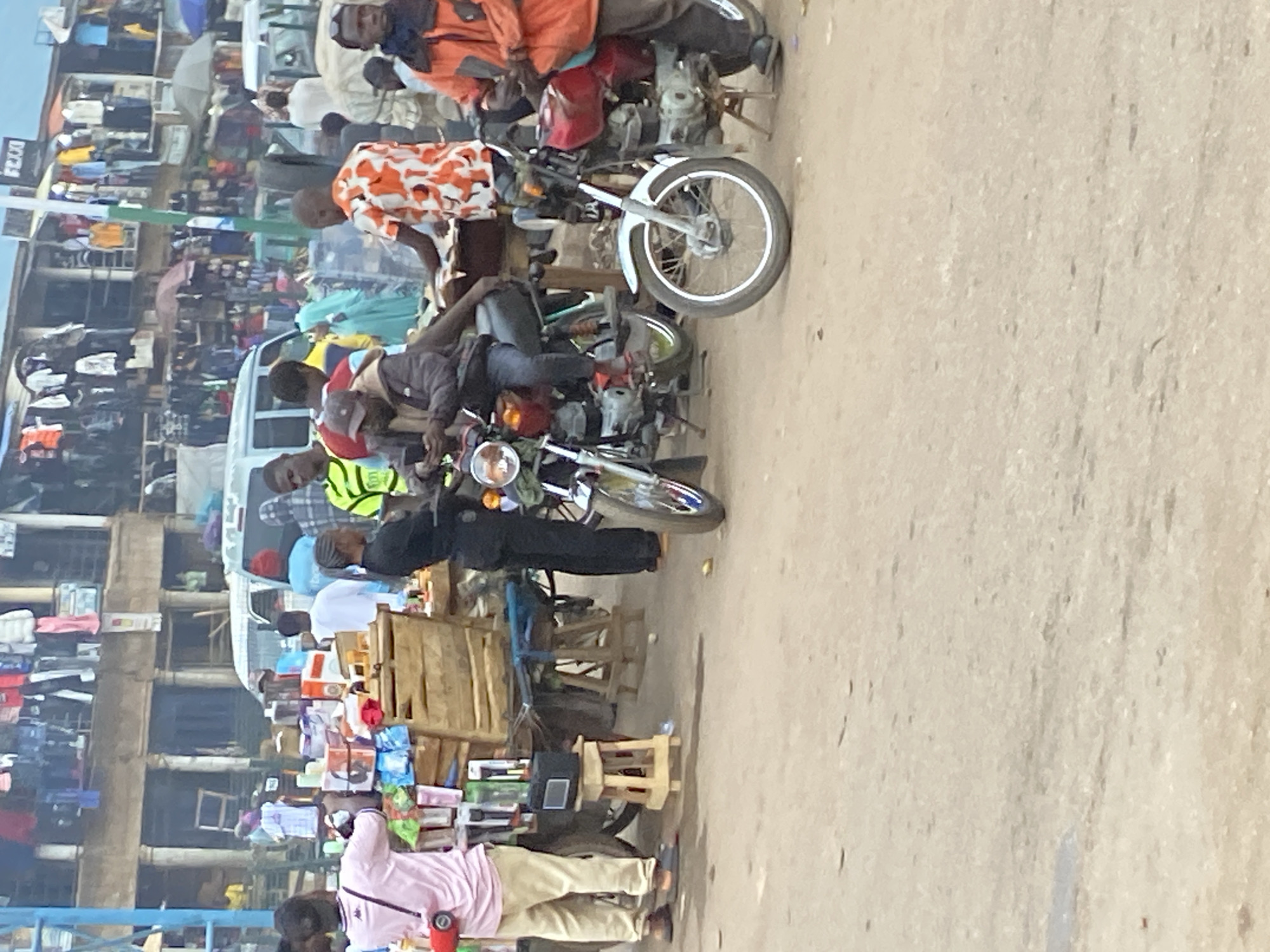 This image depicts a bustling street scene in Bukuru, Nigeria, characteristic of a market or commercial district. The photograph appears to be taken during the daytime under clear weather conditions.
The scene is densely populated with numerous people, primarily men, engaged in various activities. Many are either riding or standing near motorcycles, commonly known as "boda-bodas," which are a prominent mode of transport. One person in the midground wears a distinctive orange and white patterned shirt, while another is seen in a yellow-green reflective vest.
Along the left side of the street, multiple market stalls and makeshift shops are set up, displaying a variety of goods. These include clothing items hanging on racks and stacks of packaged products, possibly electronics or household goods, arranged on wooden tables and wooden stools. In the lower right foreground, a person in a pink shirt and khaki pants stands near one of these stalls.
The background features multi-story buildings, indicating an urban or semi-urban environment. Several white vans or cars are also visible further down the street, adding to the sense of a busy thoroughfare. The ground is an unpaved or lightly paved dirt surface.
A small white sign with the text "PZOO" is visible on a utility pole in the upper left background. The overall impression is one of vibrant daily life, with active commerce and transportation contributing to a dynamic market atmosphere.
