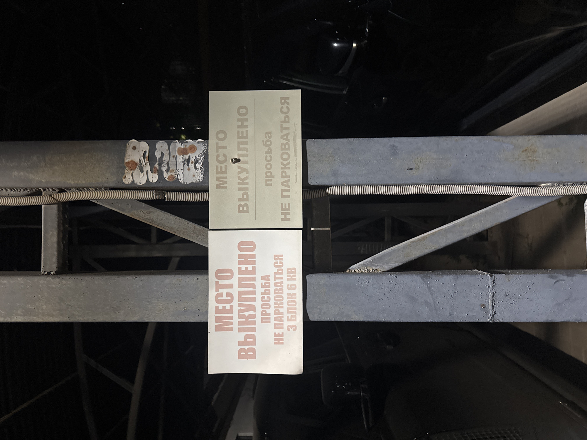 Two signs are attached to a metal pillar in what appears to be an outdoor parking area at night. The signs are white with pink text and are in Russian. The left sign reads "МЕСТО ВЫКУПЛЕНО ПРОСЬБА НЕ ПАРКОВАТЬСЯ З БЛОК 6 КВ", which translates to "PLACE BOUGHT PLEASE DO NOT PARK BLOCK 3 KV 6". The right sign reads "МЕСТО ВЫКУПЛЕНО ПРОСЬБА НЕ ПАРКОВАТЬСЯ", which translates to "PLACE BOUGHT PLEASE DO NOT PARK". There are several peeling stickers on the pillar above the signs. Two dark-colored vehicles are partially visible behind the pillar, suggesting this is a parking lot or garage. The overall lighting is dim, indicating nighttime. No people are visible in the frame. Without further context or information within the image itself, the specific location in Almaty, Kazakhstan, cannot be determined.