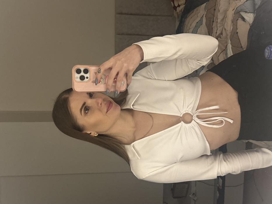 A woman takes a mirror selfie. She holds her phone, adorned with a pink case, in her right hand, capturing herself from the chest up. She wears a white, long-sleeved crop top with a circular accent and tie strings. Her dark hair is parted on the side. She appears to be standing in a room with neutral-toned walls and furniture.