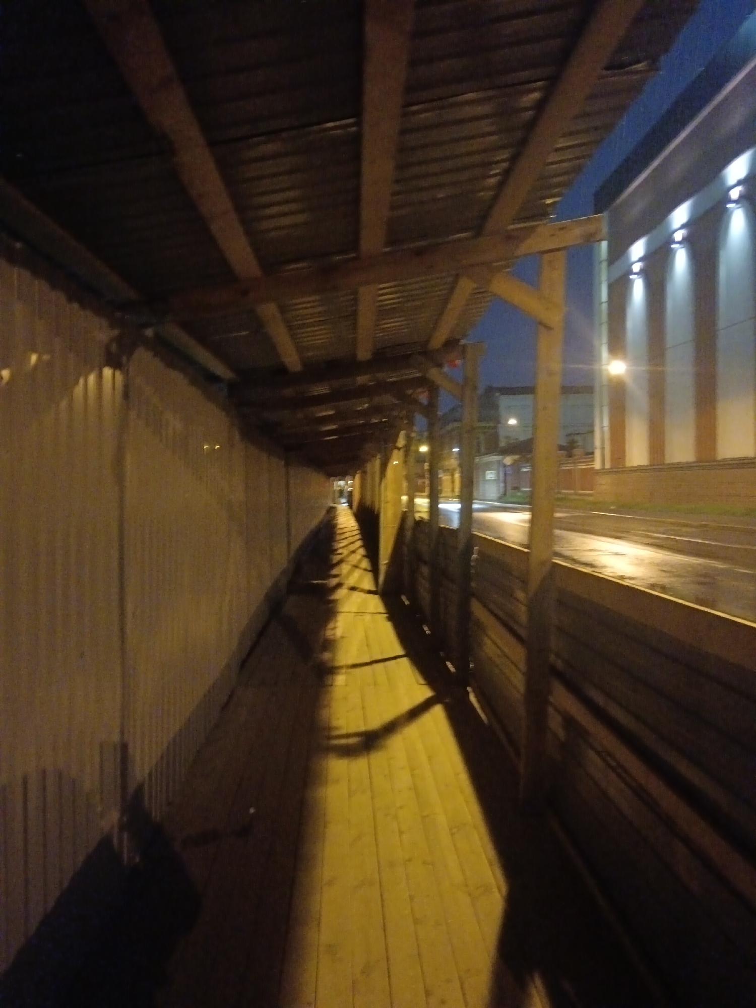 The image depicts a covered pedestrian walkway constructed with wooden beams and corrugated metal roofing. The walkway appears to be along a street or construction site at night, with artificial lighting illuminating the scene. The ground surface is made of wooden planks. On the right side of the walkway, there is a low wooden fence. In the background, a building with vertical lights is visible, and a street with wet pavement and car headlights can be seen. The overall impression is a temporary or utilitarian structure. The presence of the specific building with vertical lights and the general urban environment suggest this scene is likely located in Saint Petersburg, Russia, possibly in an area undergoing construction or renovation.