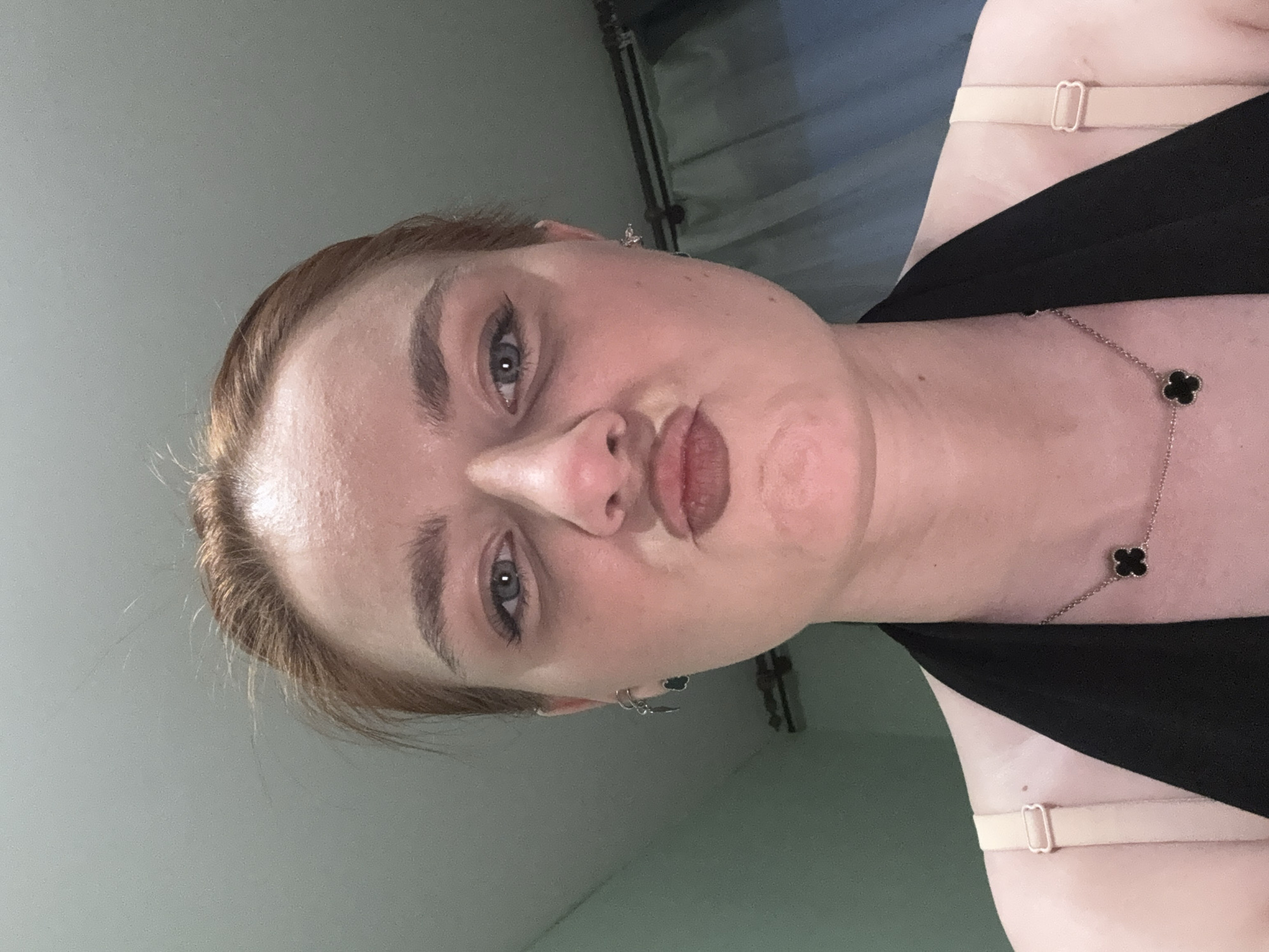 A woman with fair skin and reddish-blonde hair pulled back is looking at the camera with pursed lips. She is wearing a black top with thin straps and a delicate silver necklace with clover-shaped pendants. The background is a gradient of light blue and green, suggesting a room or interior space. The location in the city of Sinyavino, Russia is not discernible from the image.