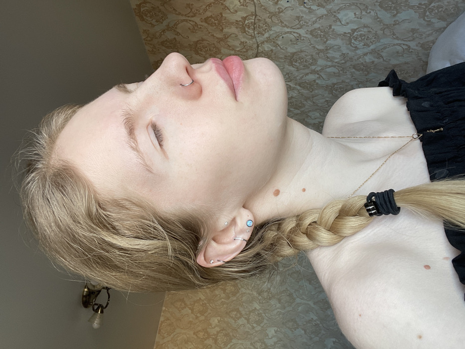 A light-skinned person with blonde hair braided and tied with black elastics, lies with closed eyes and a nose ring. The individual wears a black, ruffled top and a delicate gold necklace. Visible piercings adorn the ear, including a large opal stud and multiple rings. Several moles are apparent on the neck and shoulder. An ornate light fixture is partly visible in the background. The background has a patterned wallpaper.